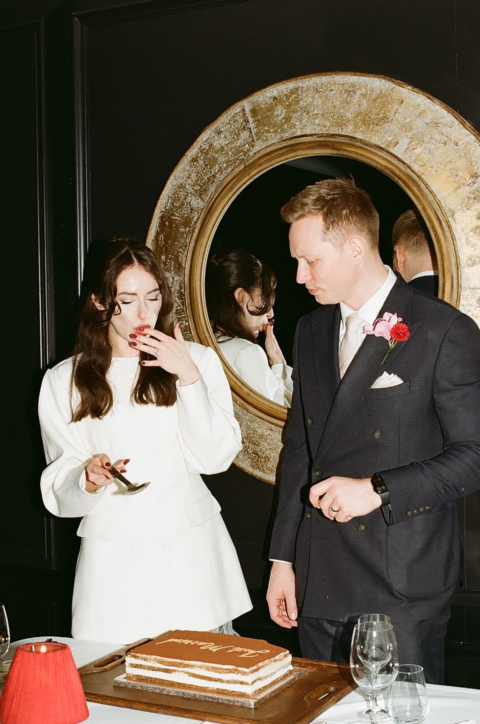 A couple in formal attire share intimate moments while looking at their phones during what appears to be a wedding reception.