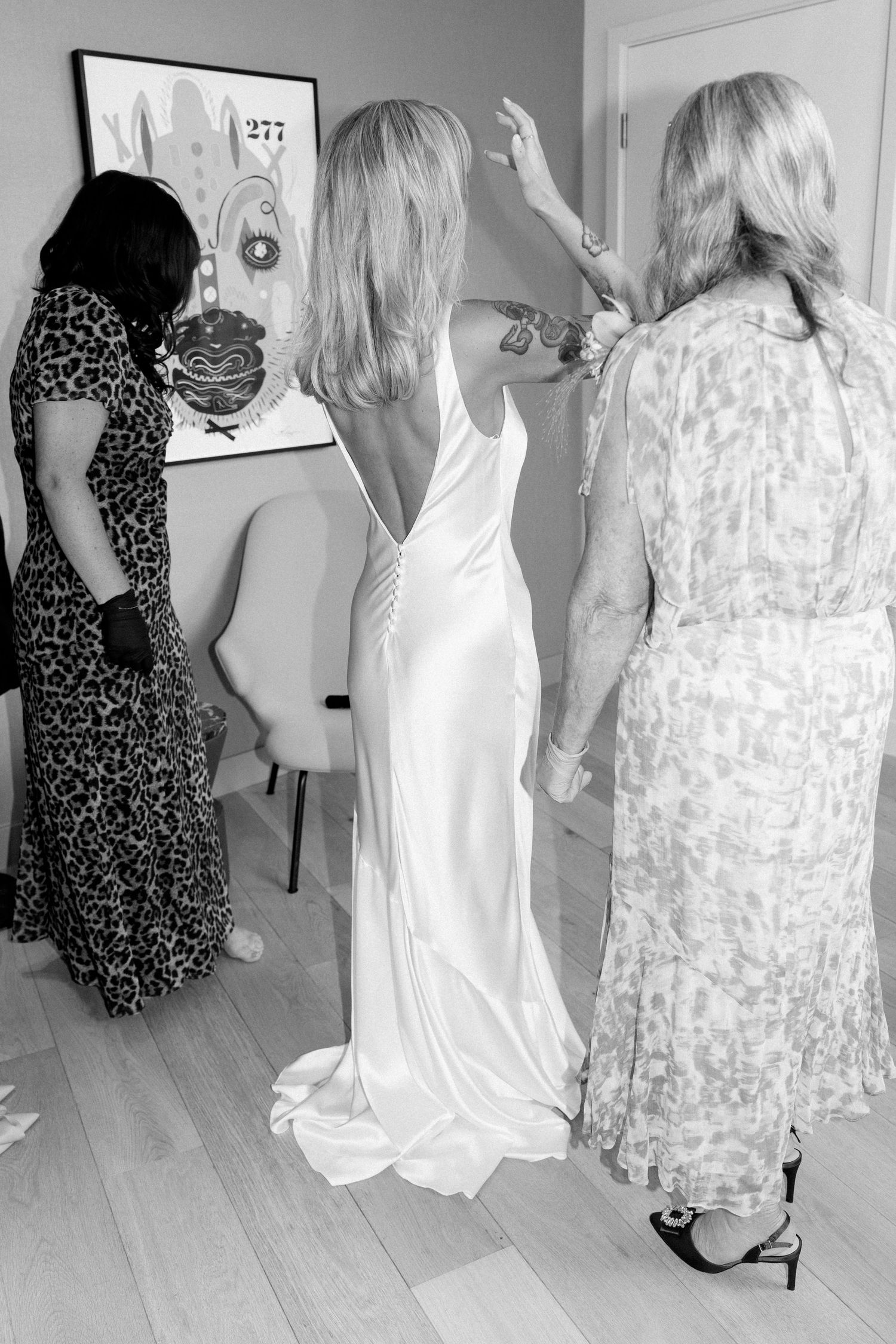 Three women getting ready in a room, one wearing a flowing white wedding dress.