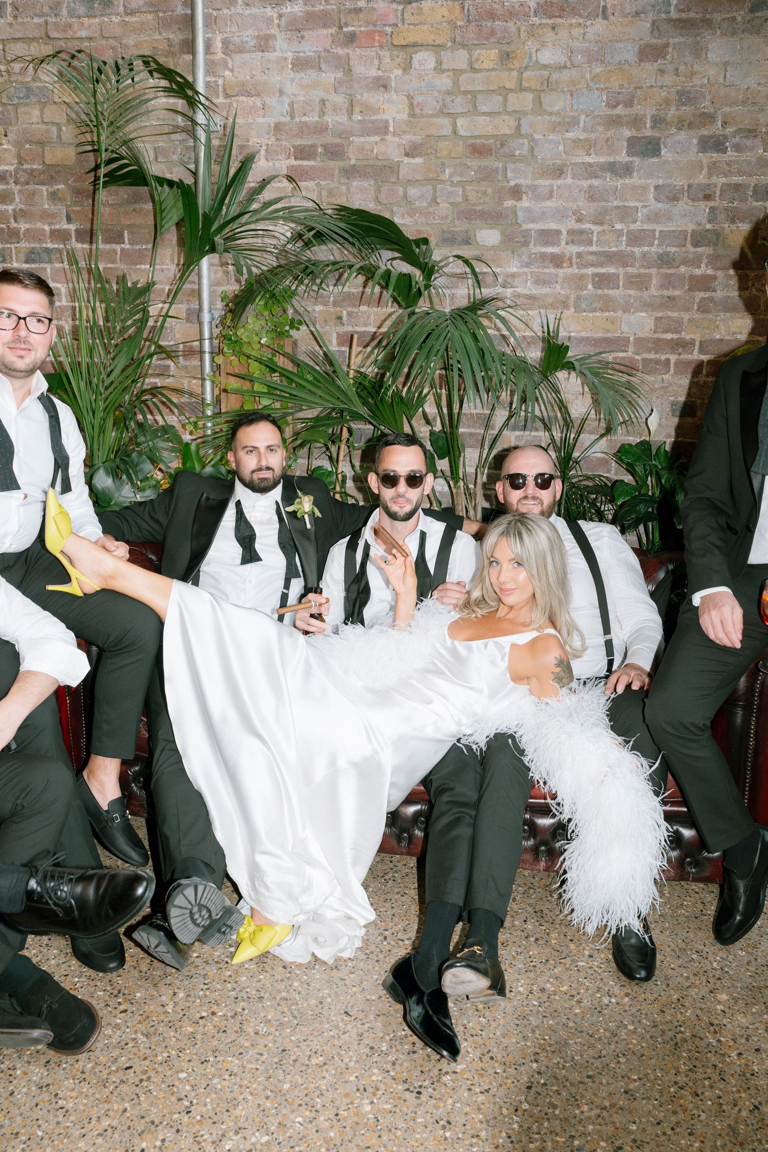 Wedding party in formal attire sitting casually against a brick wall with tropical plants.