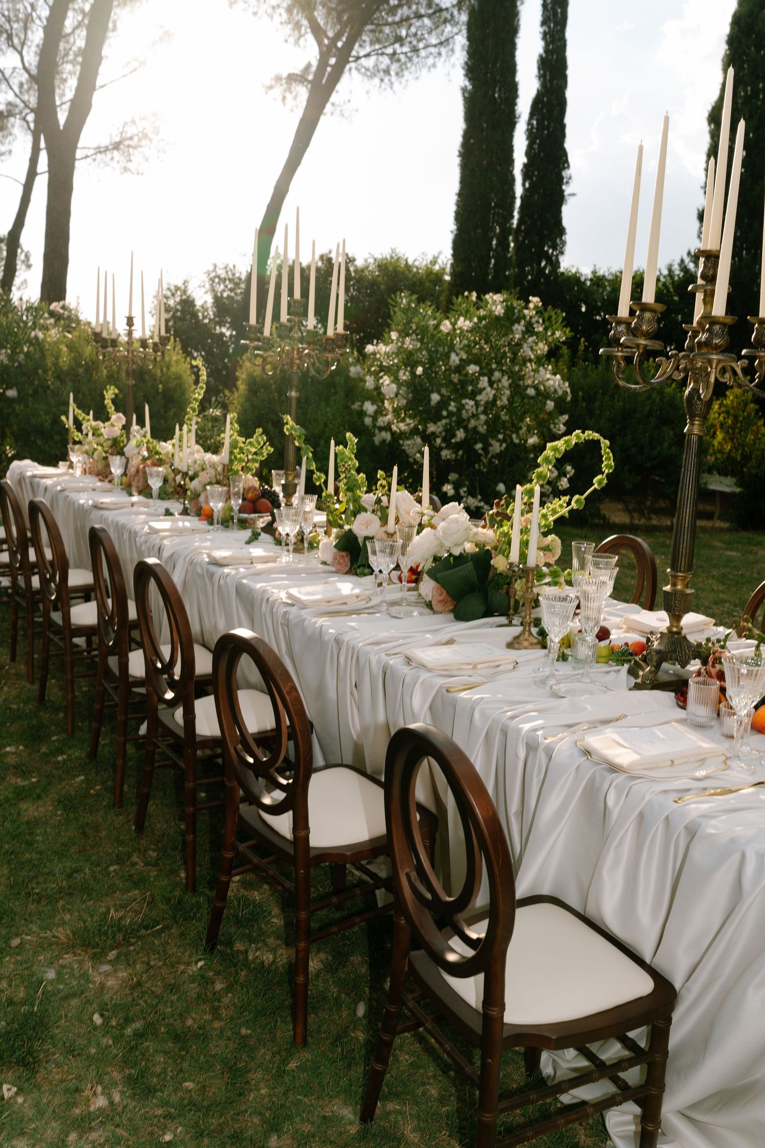 An elegant wedding table setting with white linens and floral arrangements under tall cypress trees.