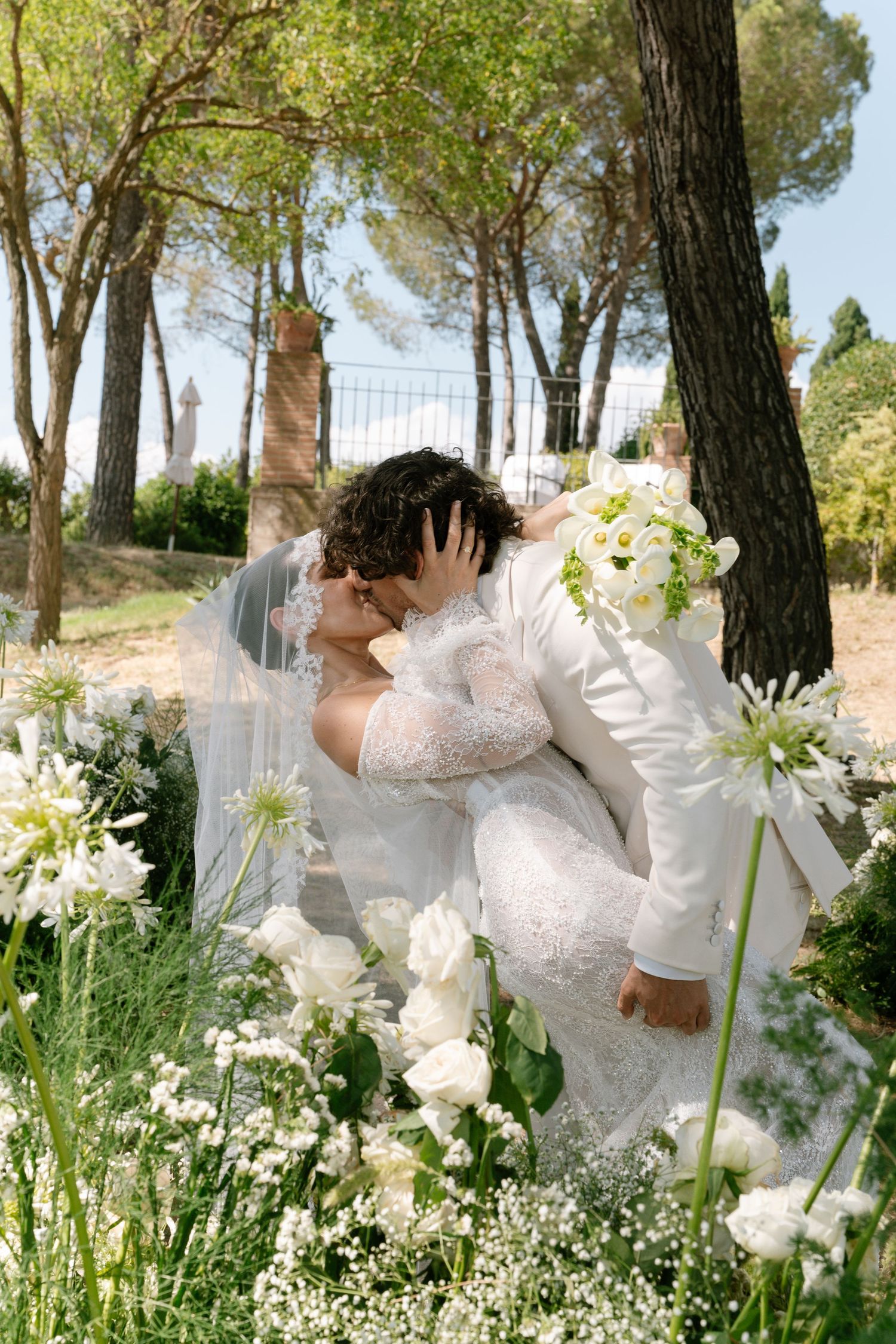 White lilies and delicate flowers create an ethereal garden scene under tall pine trees.