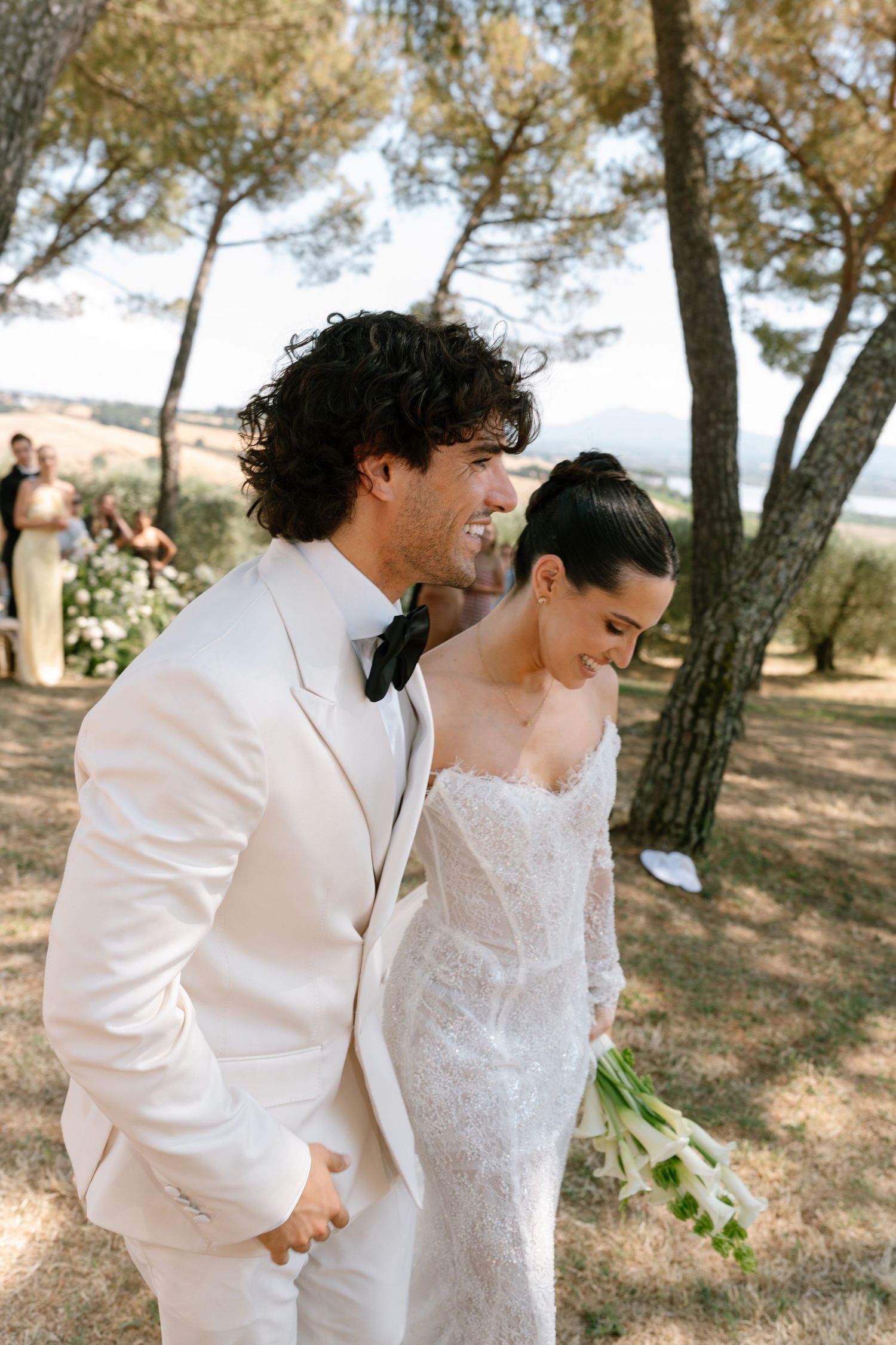 Couple in white wedding attire walking through a sun-dappled grove.