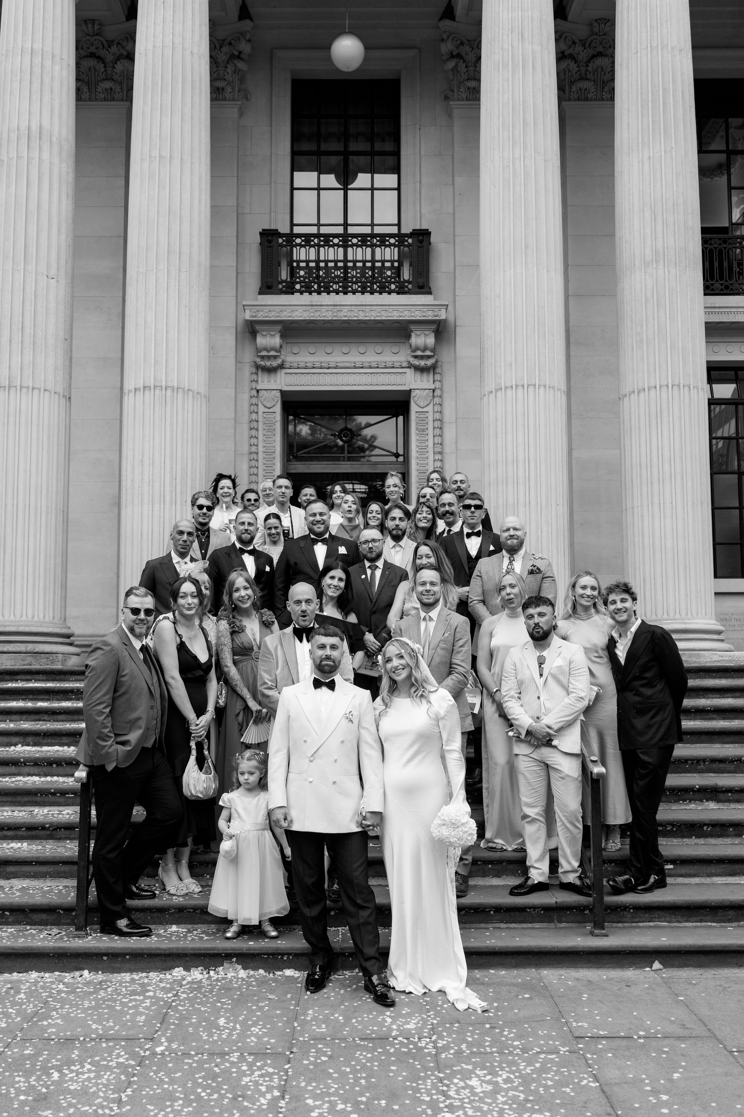 Large wedding party group photo on grand stone steps of historic building.