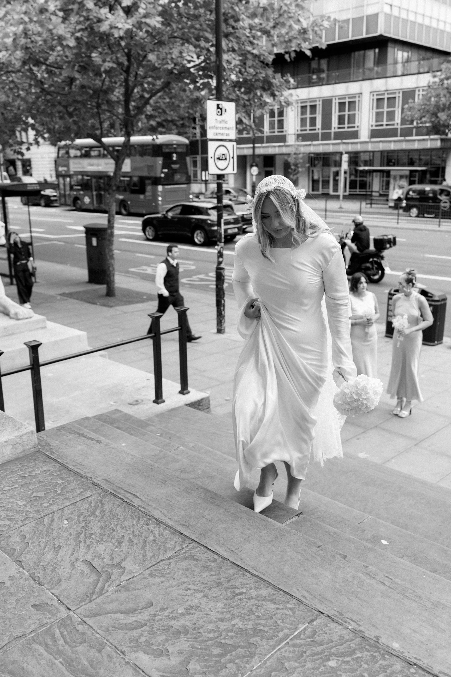 A person in a flowing white dress walks on a city street in a black and white photograph.