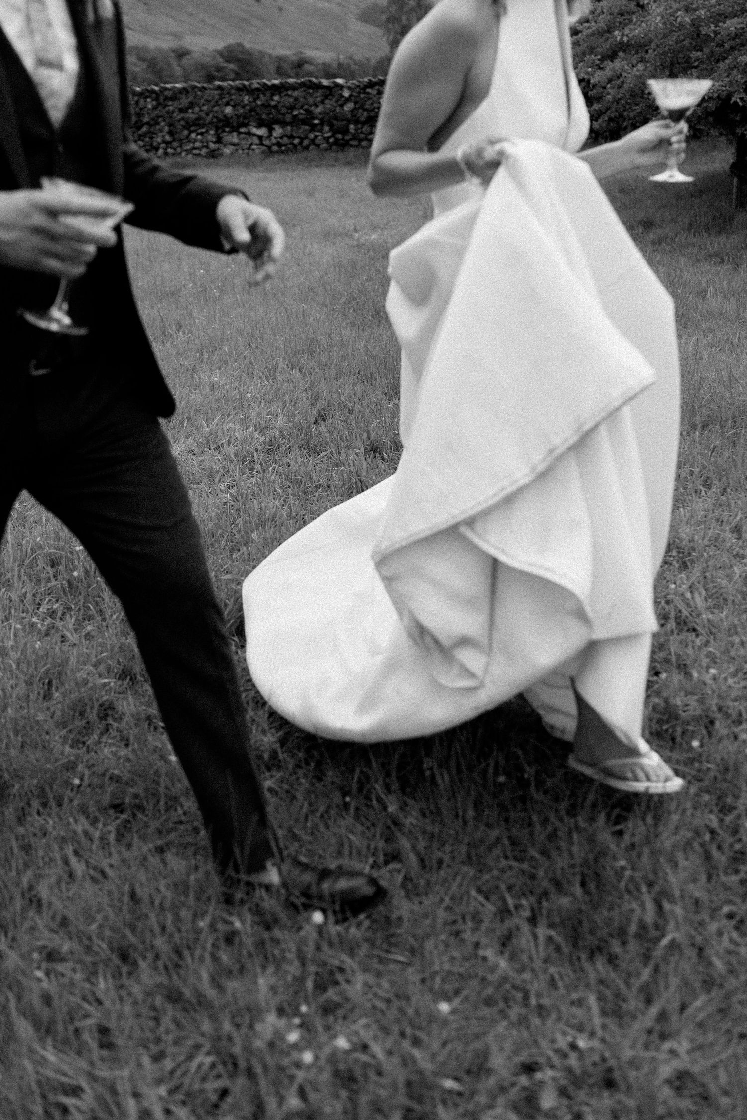 Black and white detail shot of flowing wedding dress and formal attire during outdoor celebration.