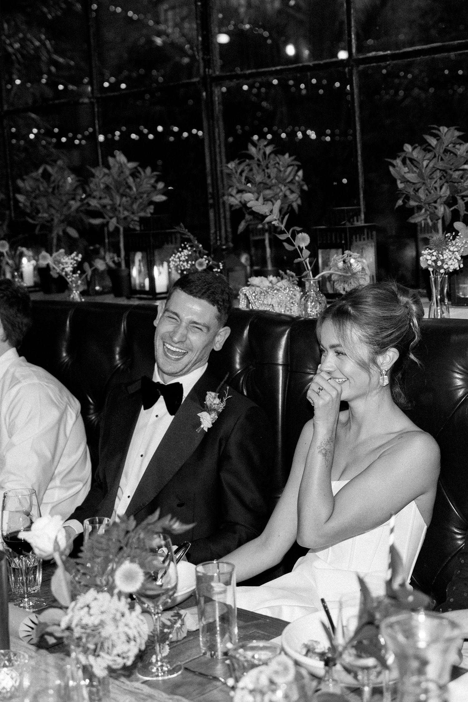 A black and white photo of a couple laughing together at an elegant reception dinner with floral centerpieces.