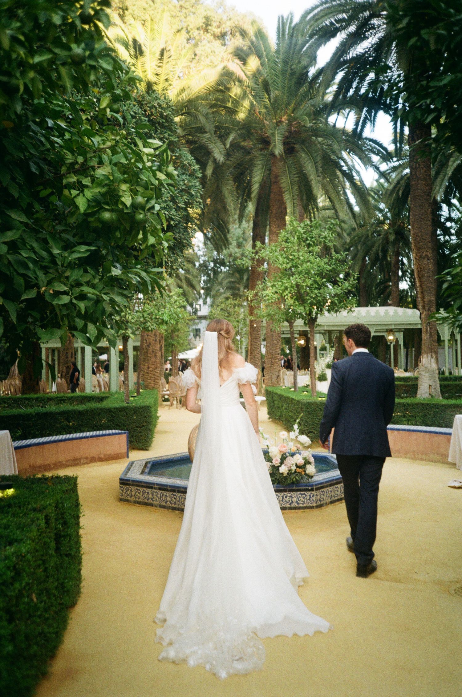 A garden pathway lined with palm trees and greenery features ornate tile borders and sandy ground.