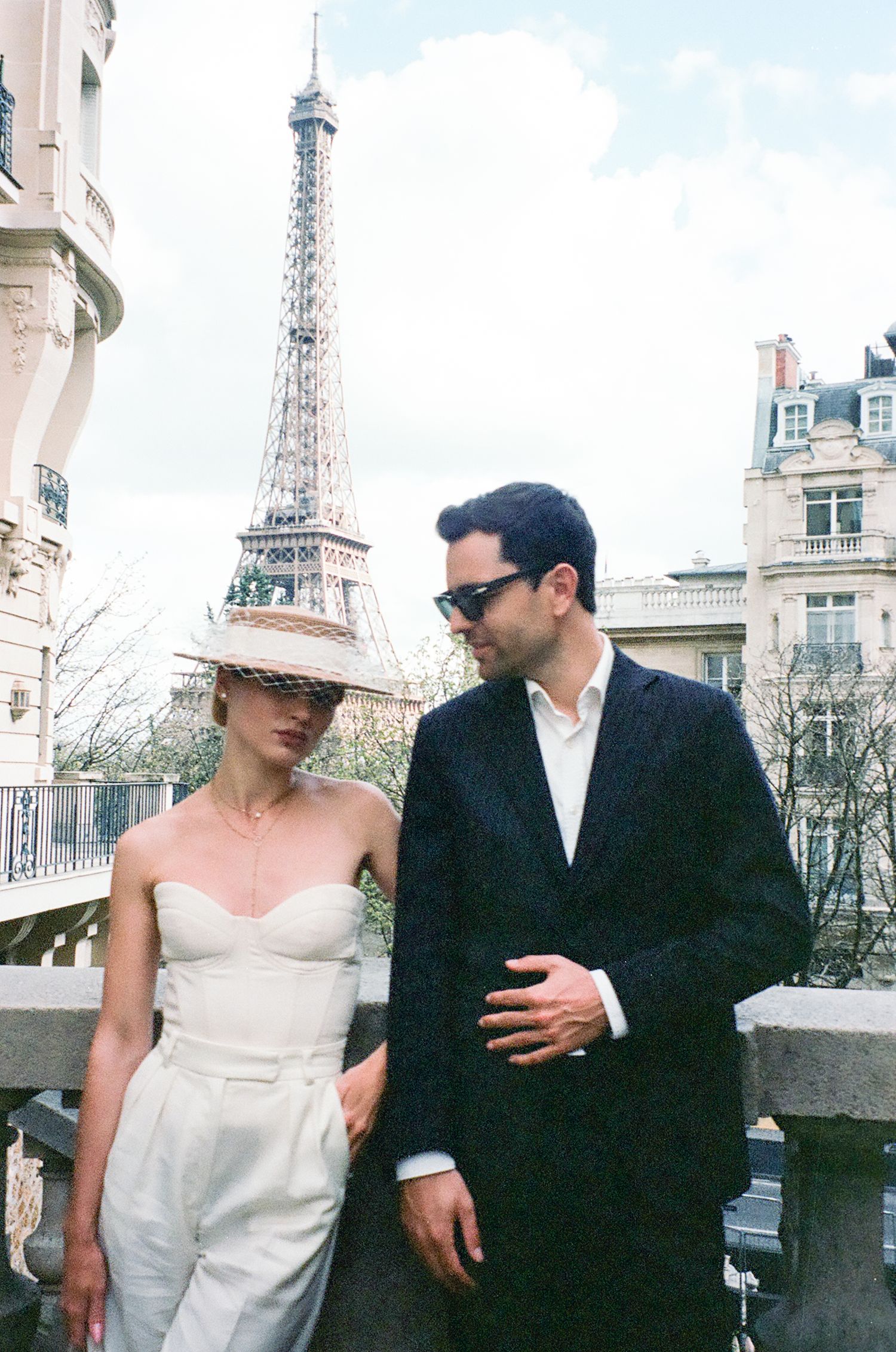A couple stands together on a balcony in Paris with the iconic Eiffel Tower visible in the background.