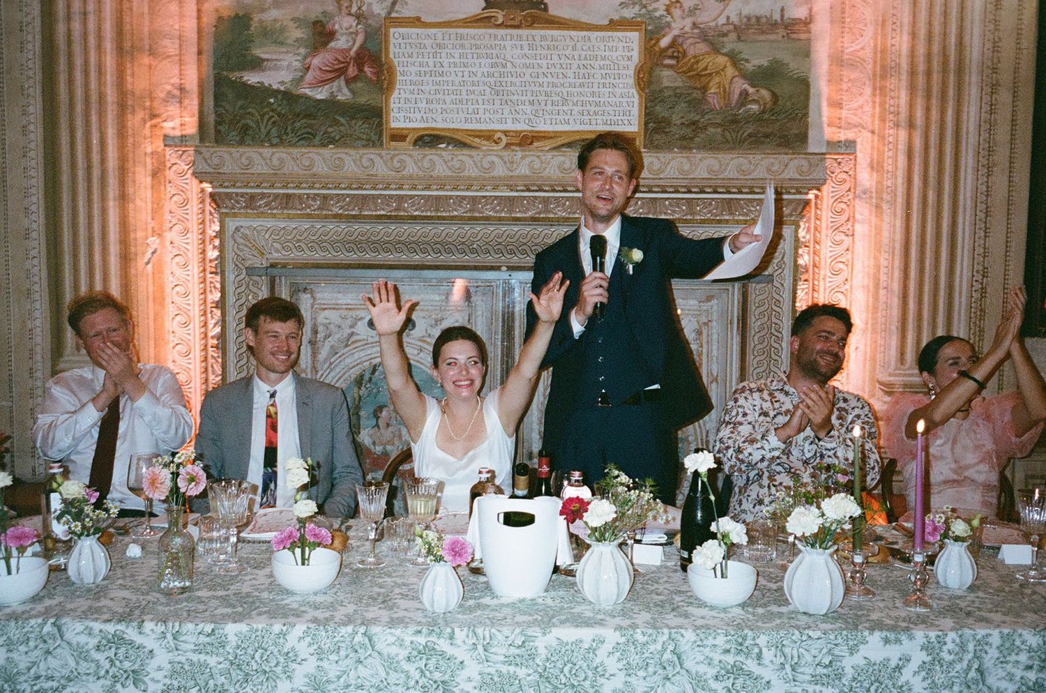 Wedding guests seated at a long banquet table in an ornate dining room raise their glasses in celebration.
