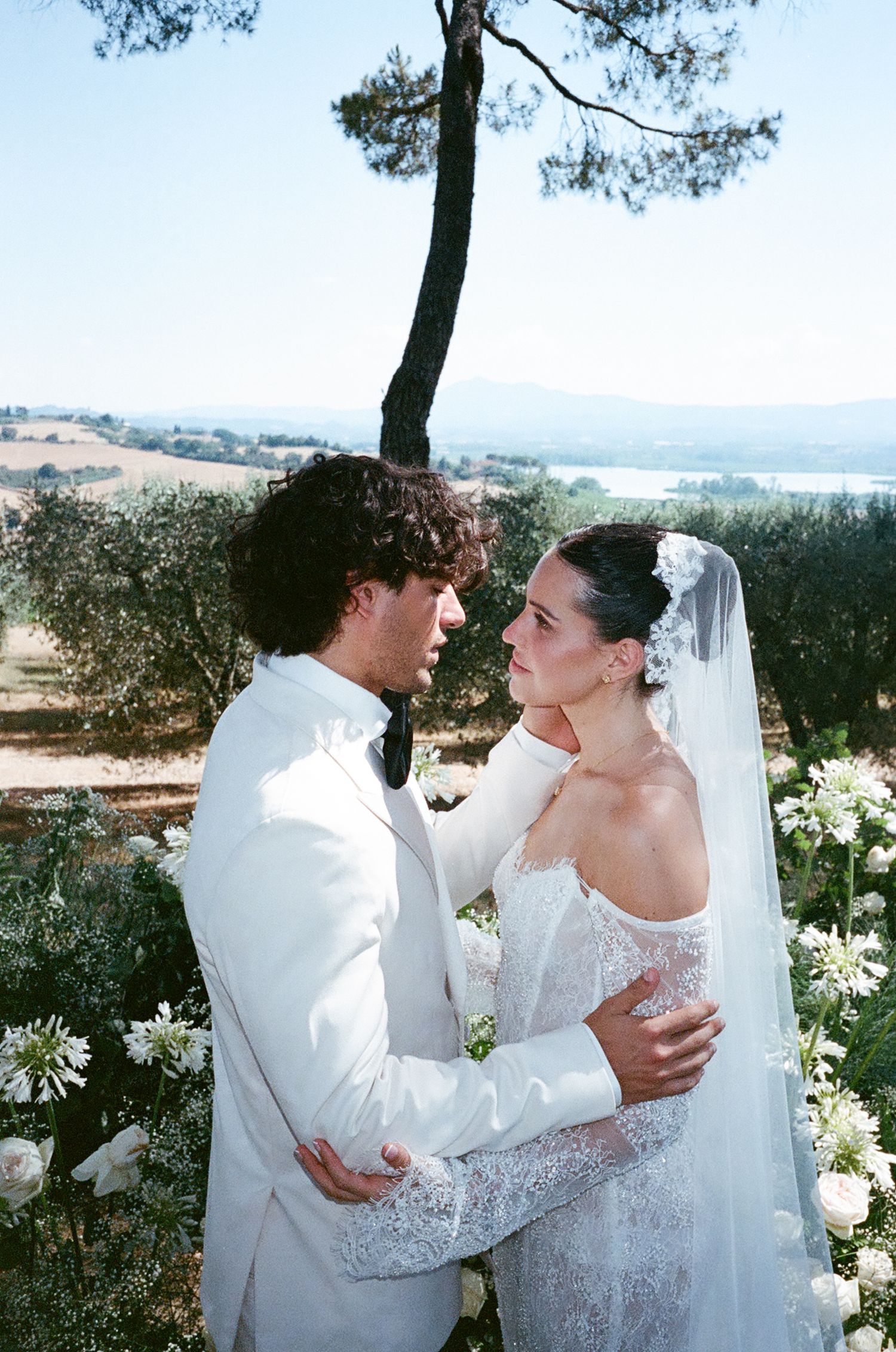 A couple embraces outdoors on their wedding day, with a scenic landscape of rolling hills and a lake in the background.