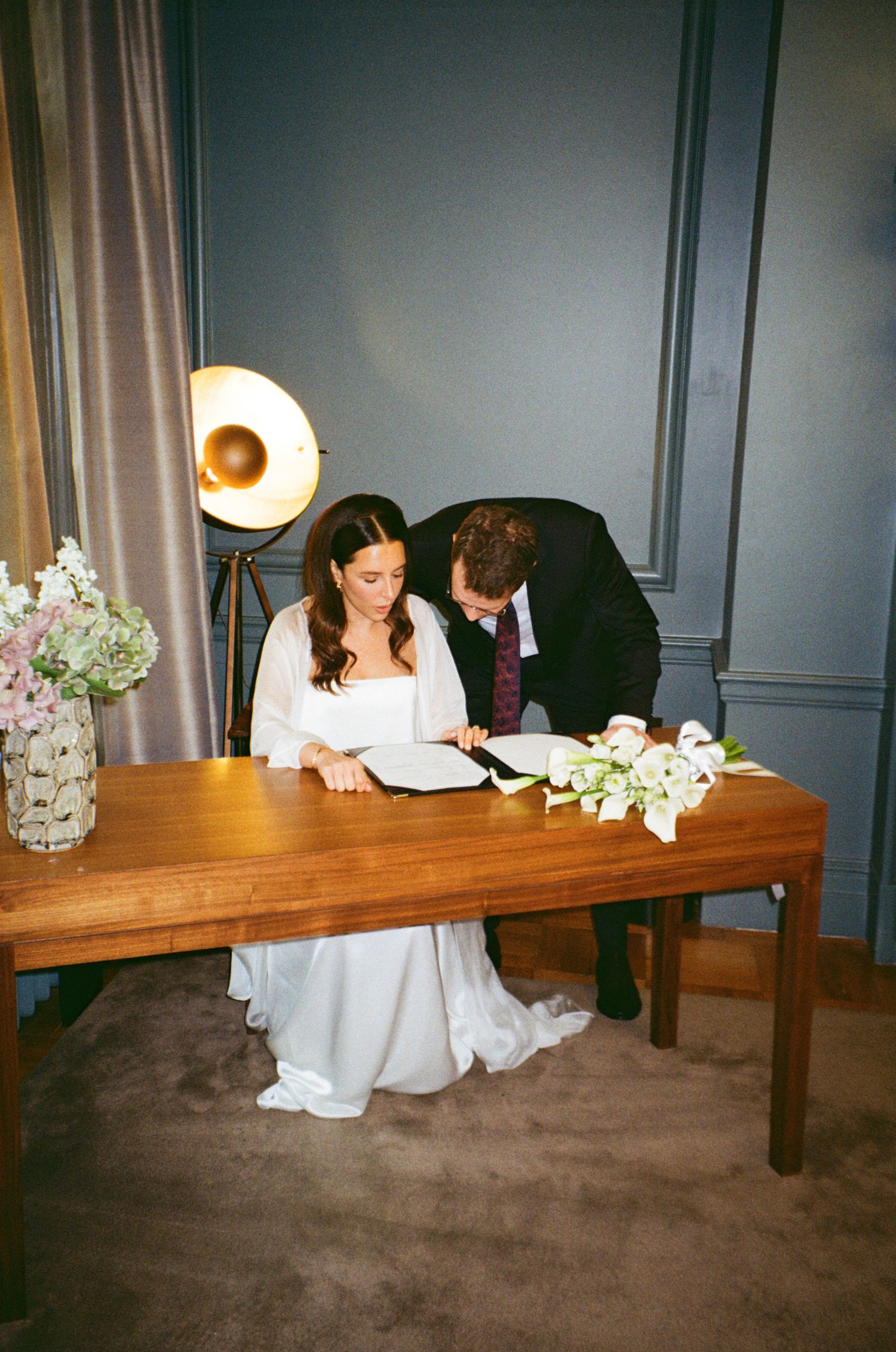 A couple in wedding attire sits together at a wooden table decorated with white flowers in an elegant room.