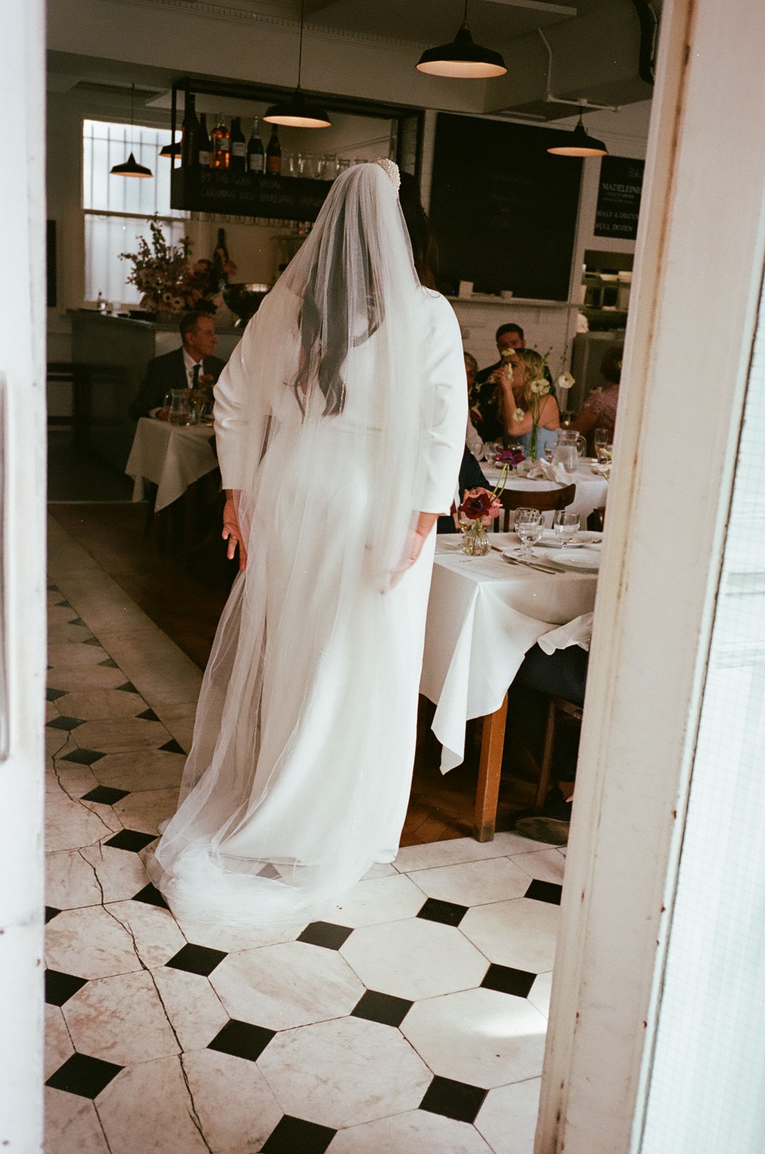 People in white wedding dresses walk across a black and white checkered floor in a bright indoor venue.