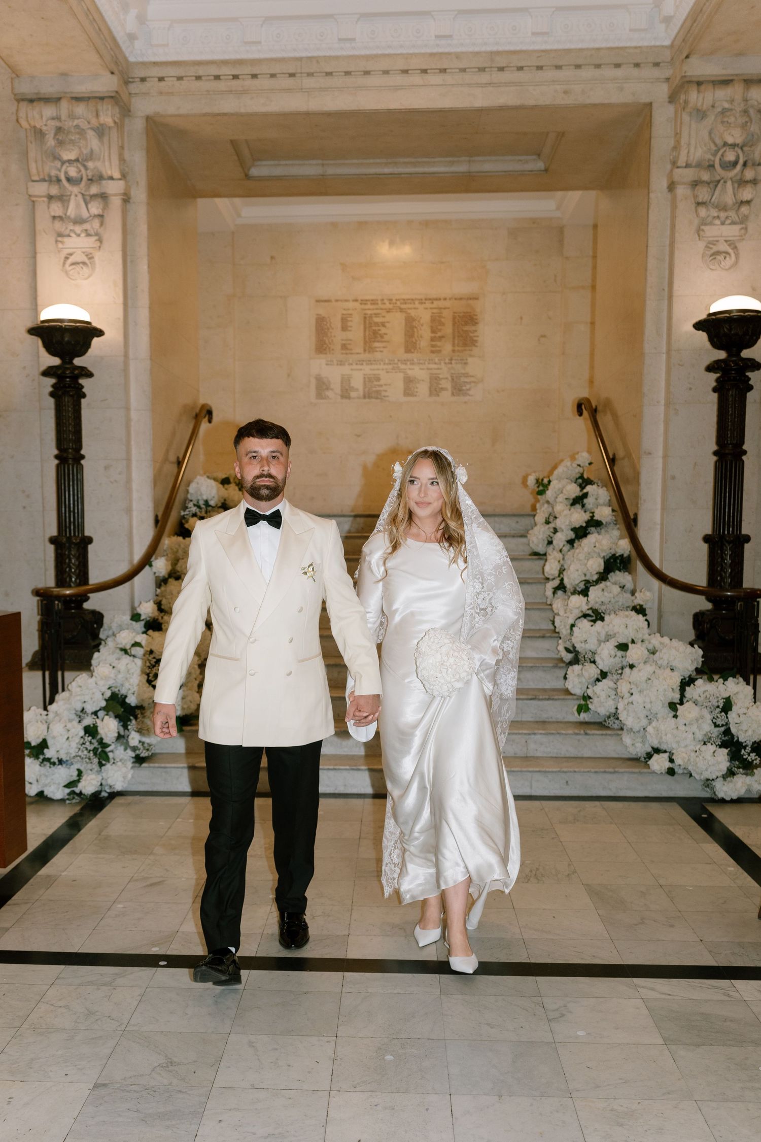 Elegant couple walks down marble hallway decorated with ornate black lamp posts and white floral arrangements.