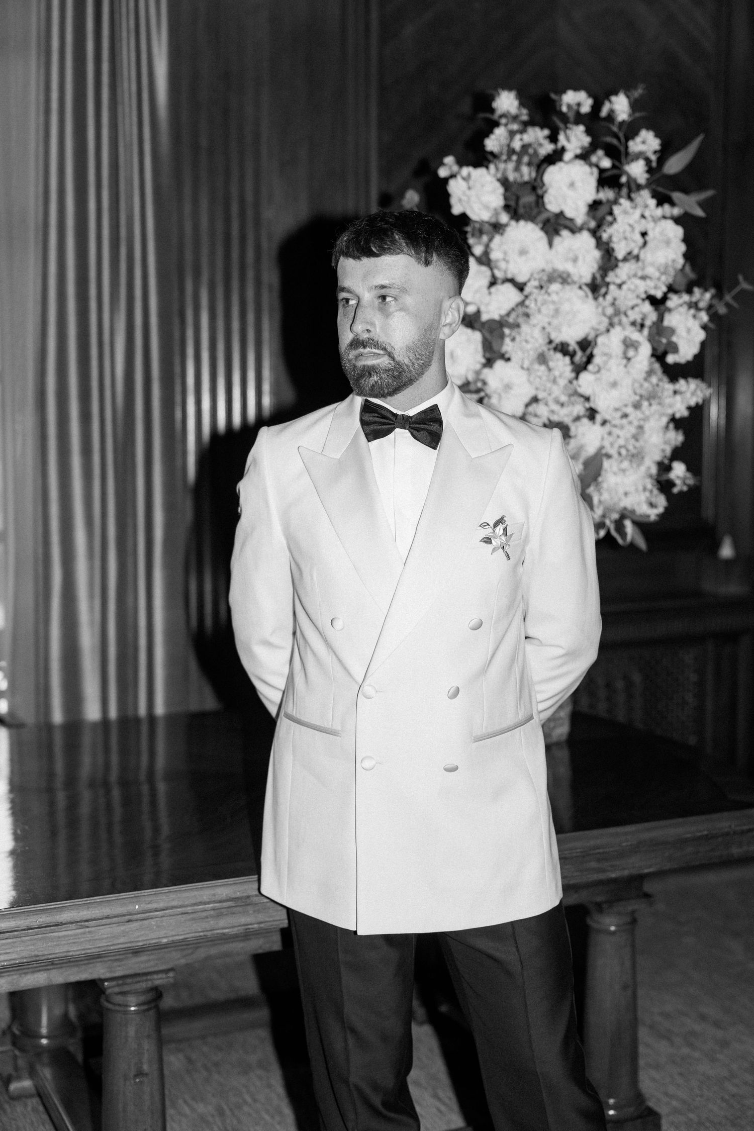Couple in formal white attire and black bow ties pose in an elegant indoor setting with floral arrangements.