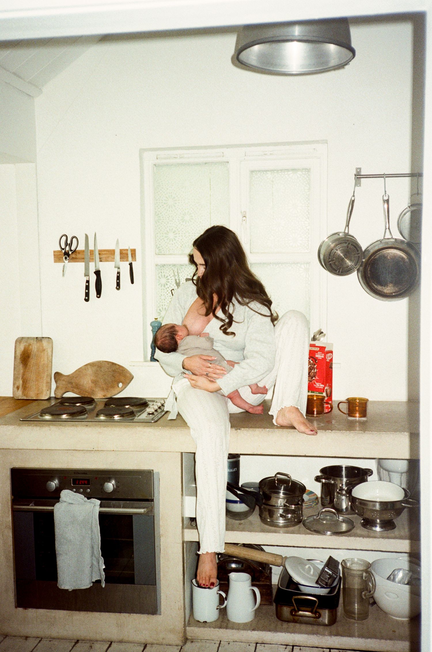 Woman sitting on kitchen counter breastfeeding a newborn baby, surrounded by pots, pans, and cooking utensils.
