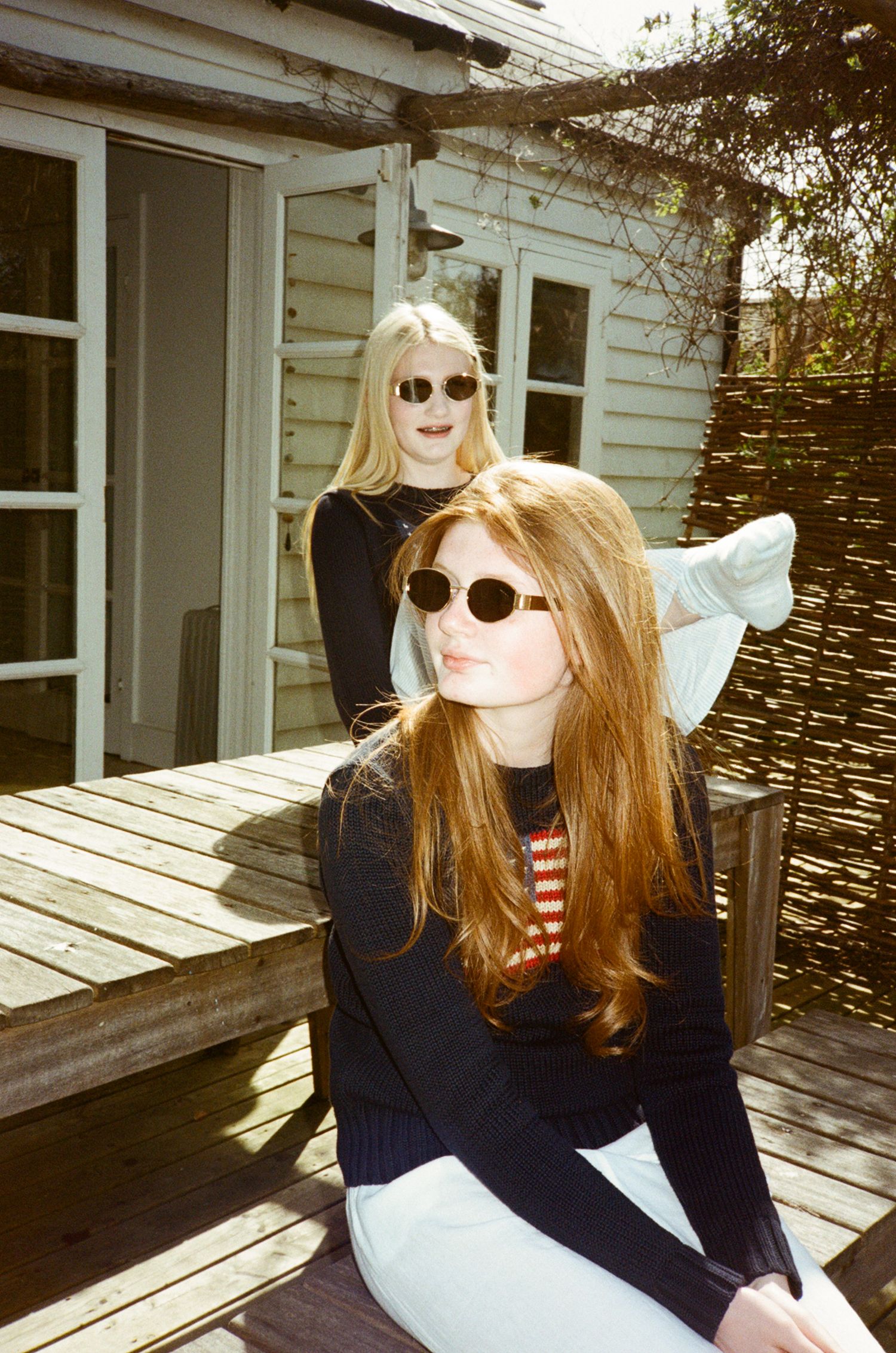 Two women in black tops on a sunny wooden deck, one wearing round sunglasses and a striped shirt.