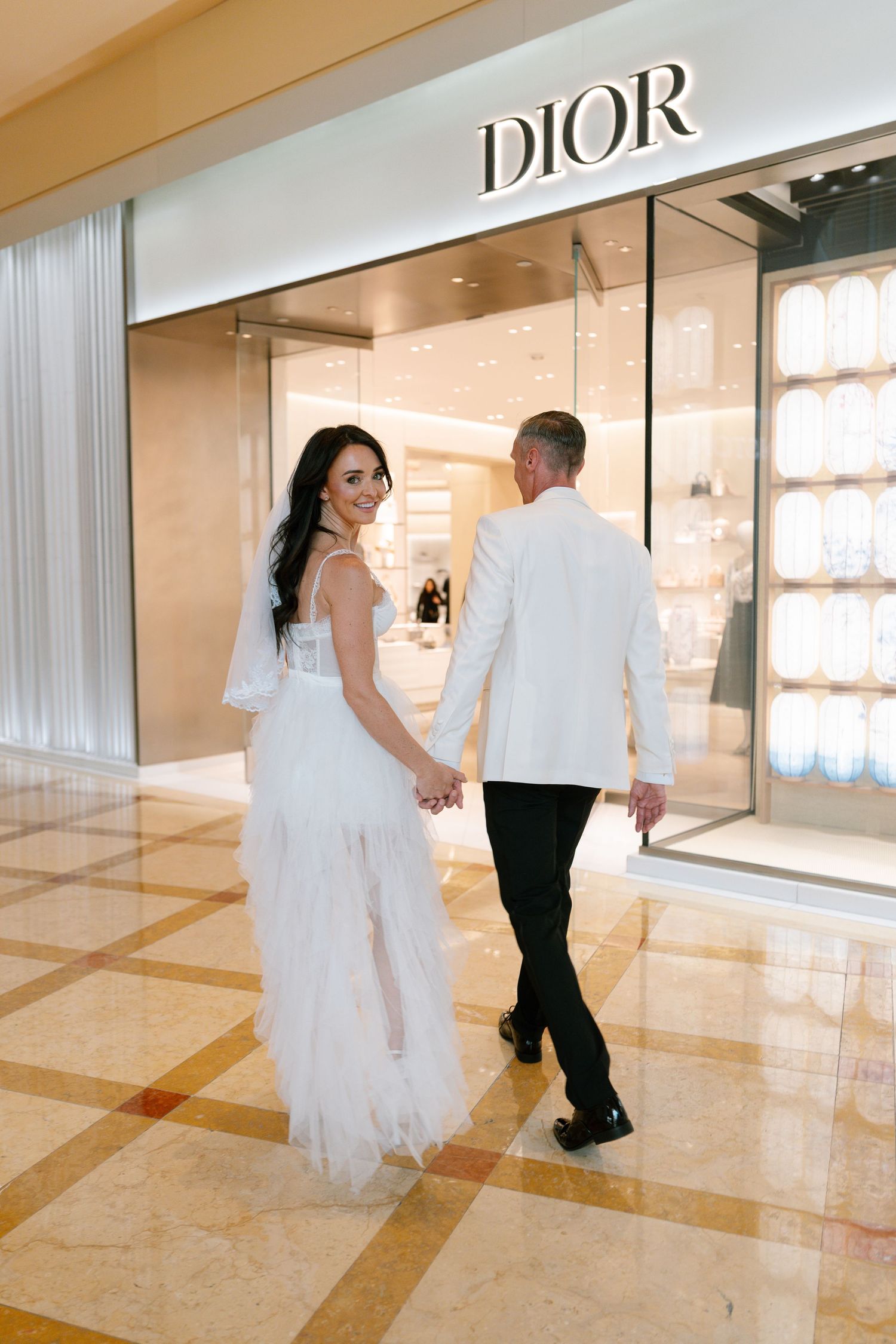 Newlyweds strolling past the Dior boutique in a luxury shopping center.
