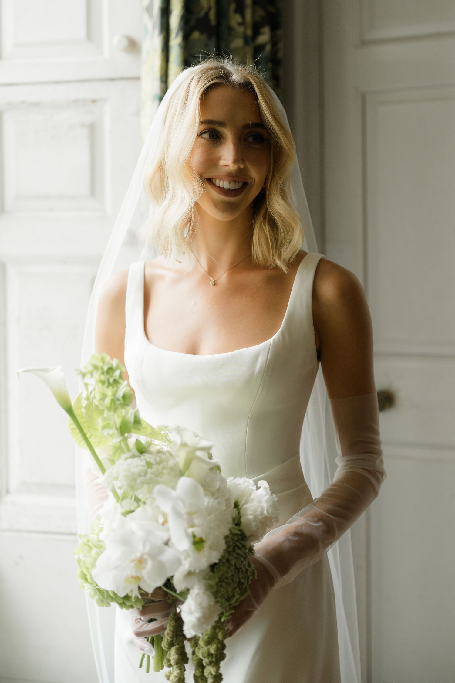 A smiling blonde bride in a white square-neck gown holds a white and green bouquet wearing a veil.