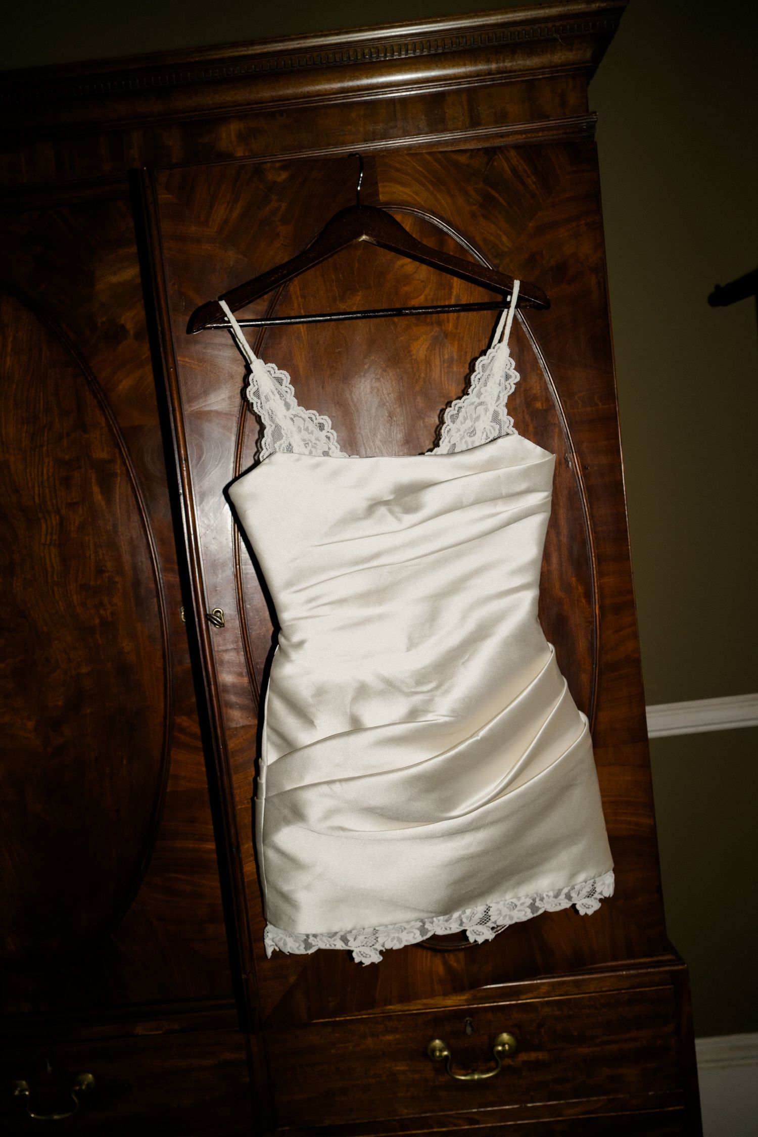 Elegant white satin and lace mini wedding dress hanging on a wooden armoire hanger.
