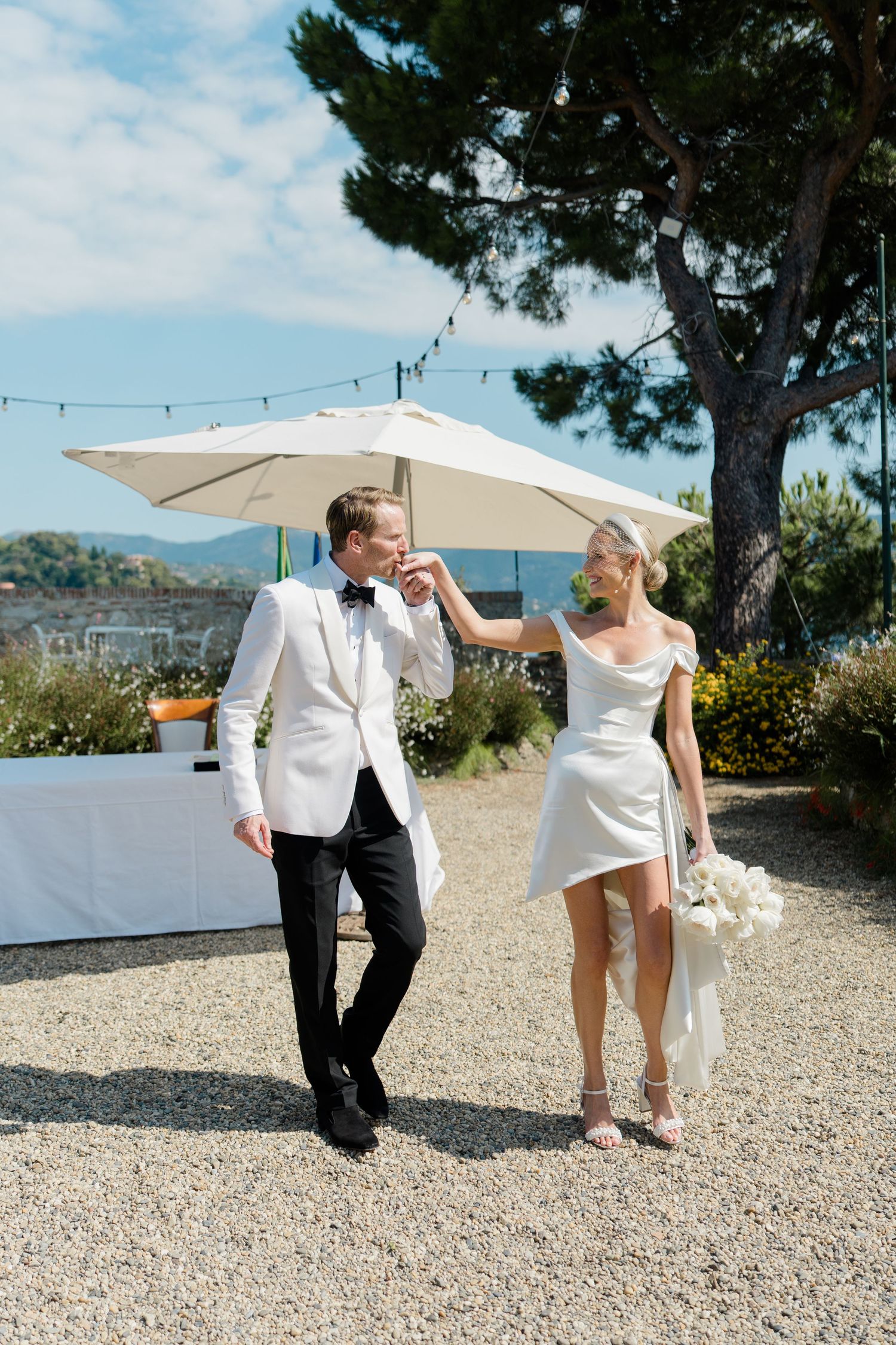 Elegant bride and groom in white outfits walk outdoors under umbrella with scenic Mediterranean view.