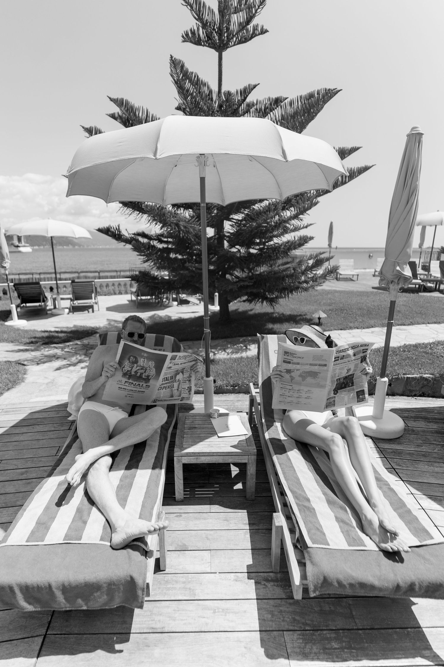 Couple relaxing on striped sun loungers reading newspapers under a beach umbrella in black and white photo.