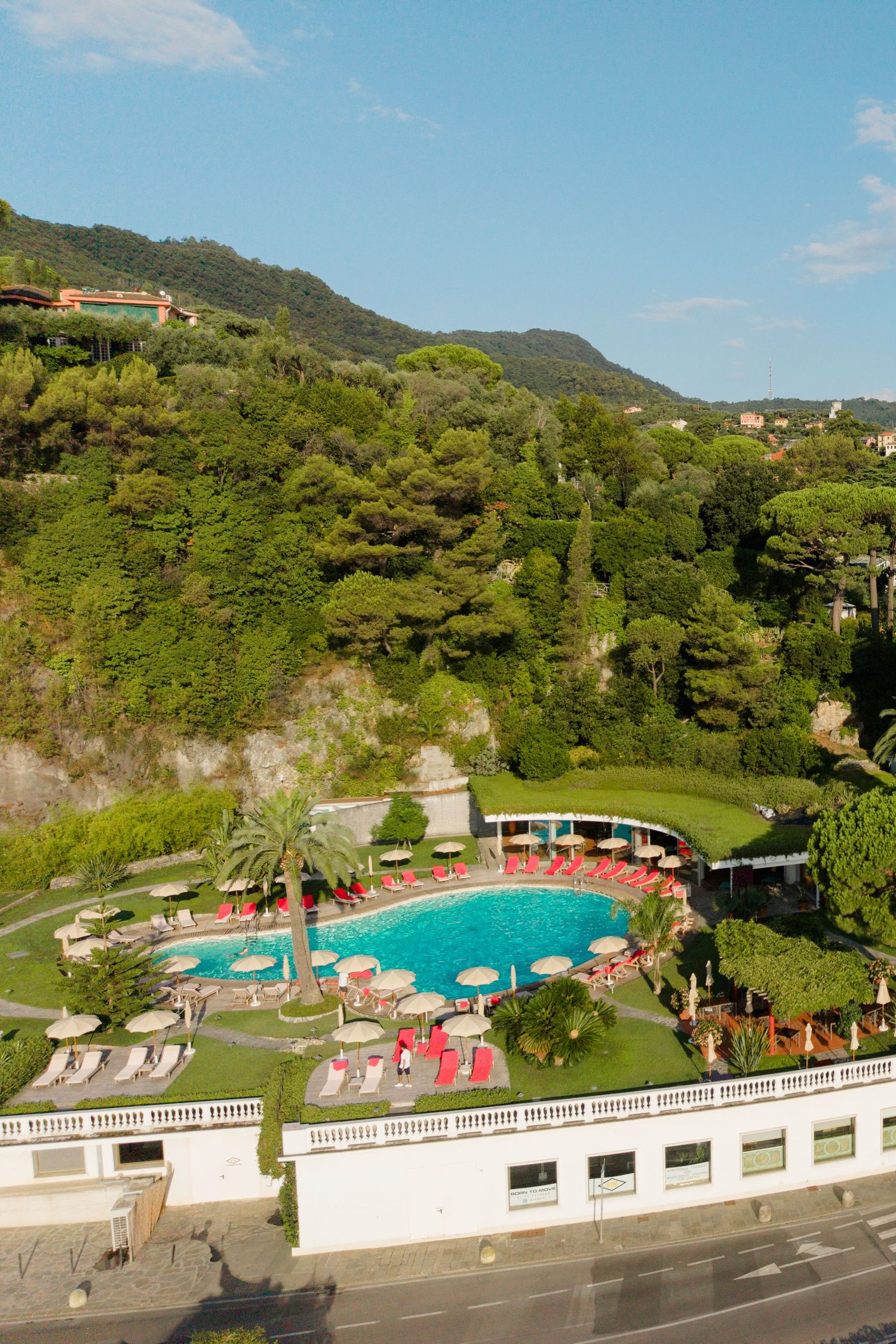 Aerial view of a luxury hotel pool surrounded by red umbrellas and lush green hills in the Italian Riviera.