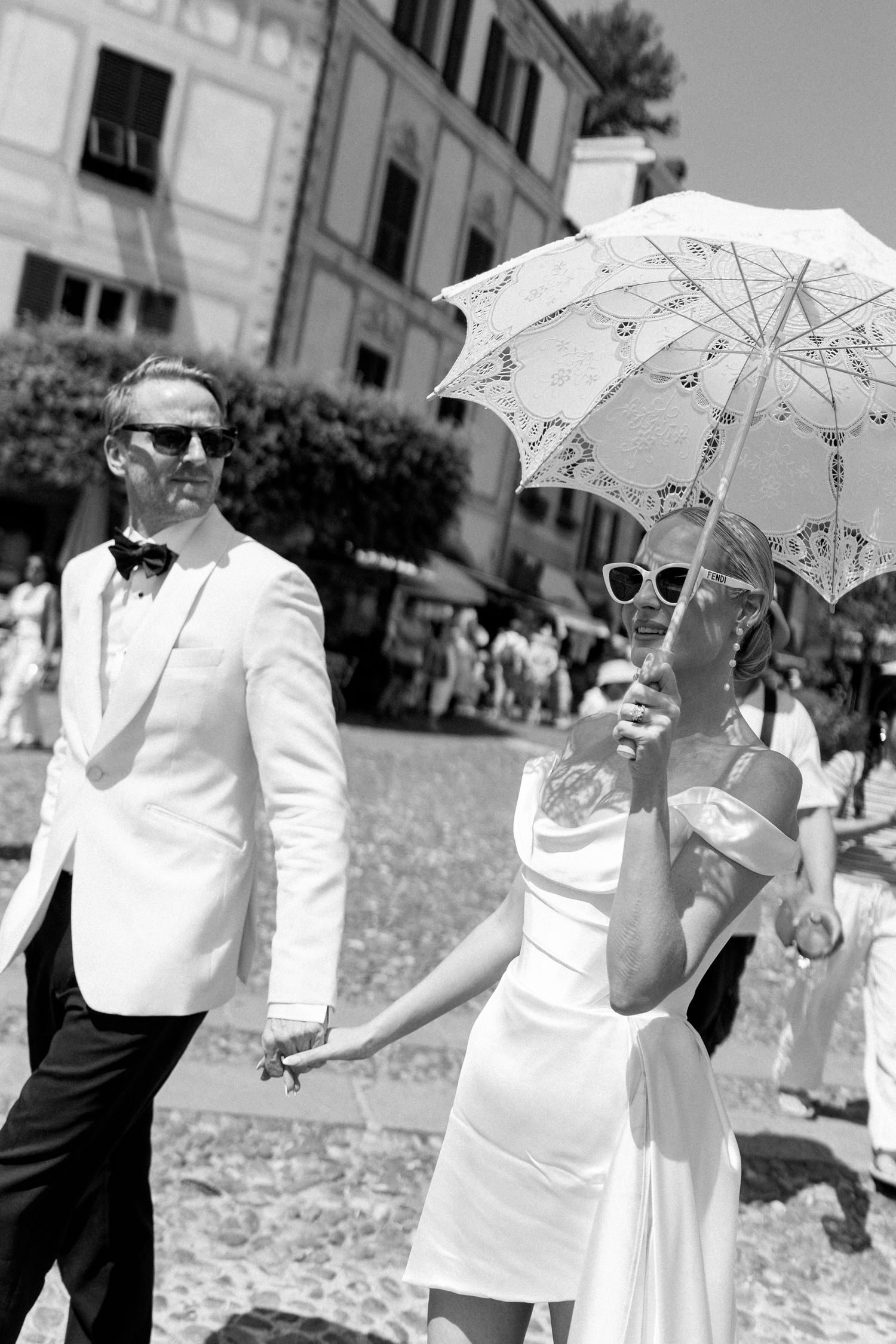 Elegant couple in white wedding attire walk hand-in-hand through sunny Italian piazza, bride holding lace parasol.