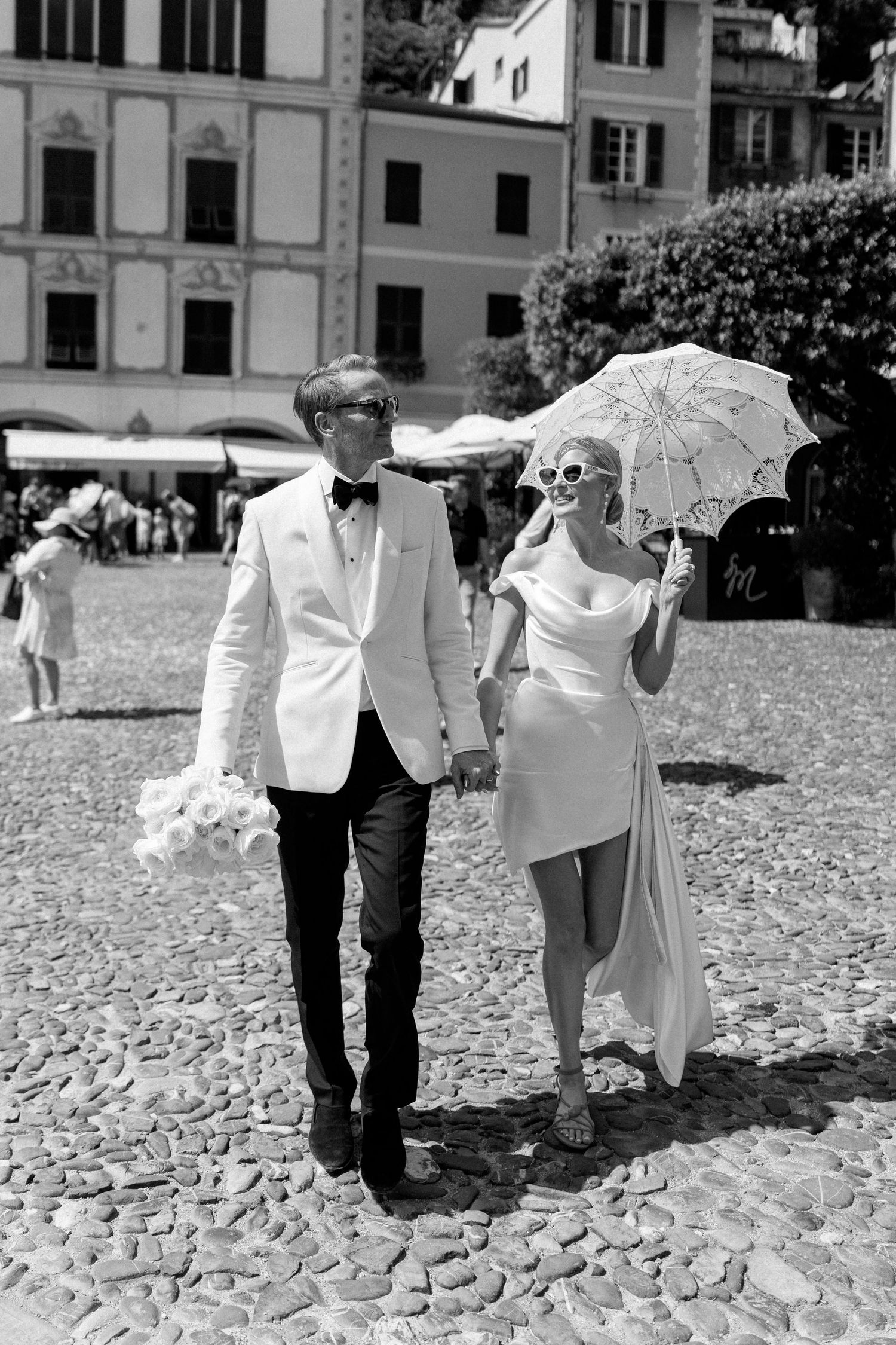 Bride and groom walk through sunny cobblestone plaza, bride holds lace parasol and sunglasses.