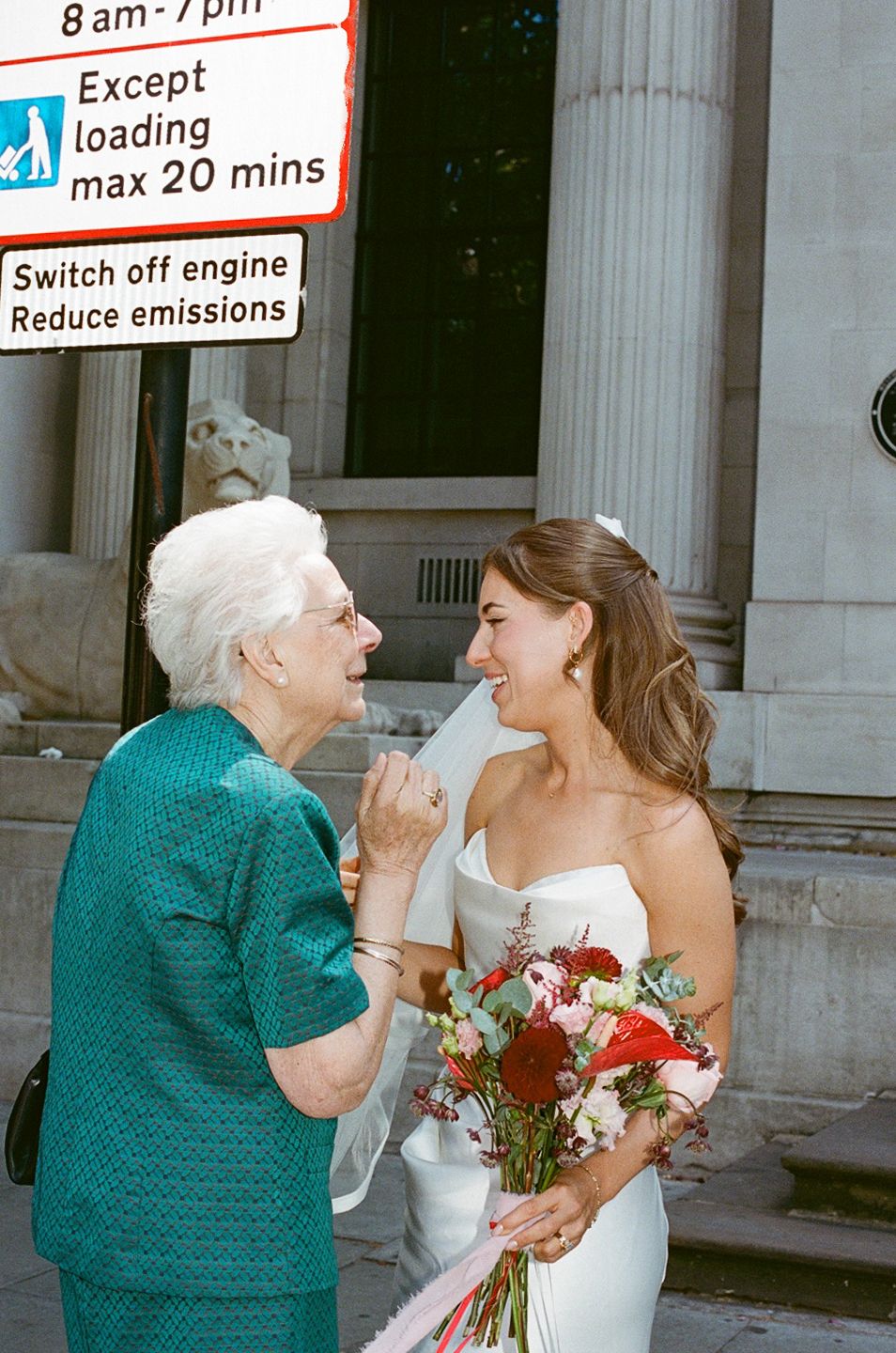 Joyful bride in white gown shares heartfelt moment with elderly woman on city street.
