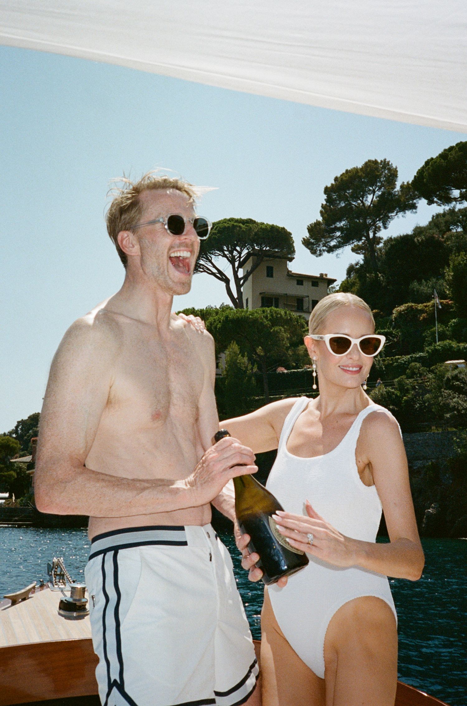 A laughing couple in swimwear on a boat, holding a champagne bottle, with Mediterranean scenery behind them.