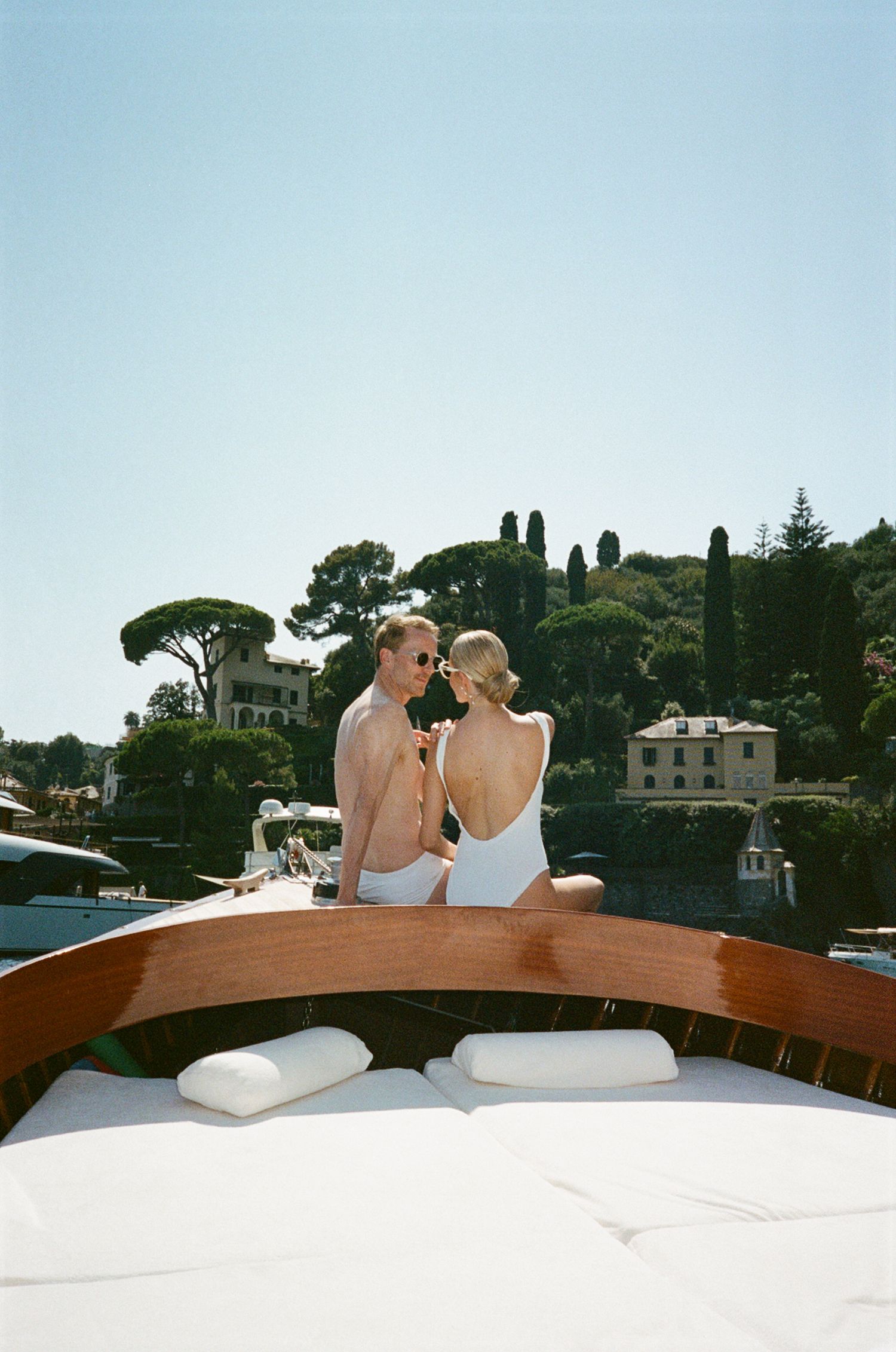 Couple in white swimwear share intimate moment on luxury wooden boat near Italian villa.