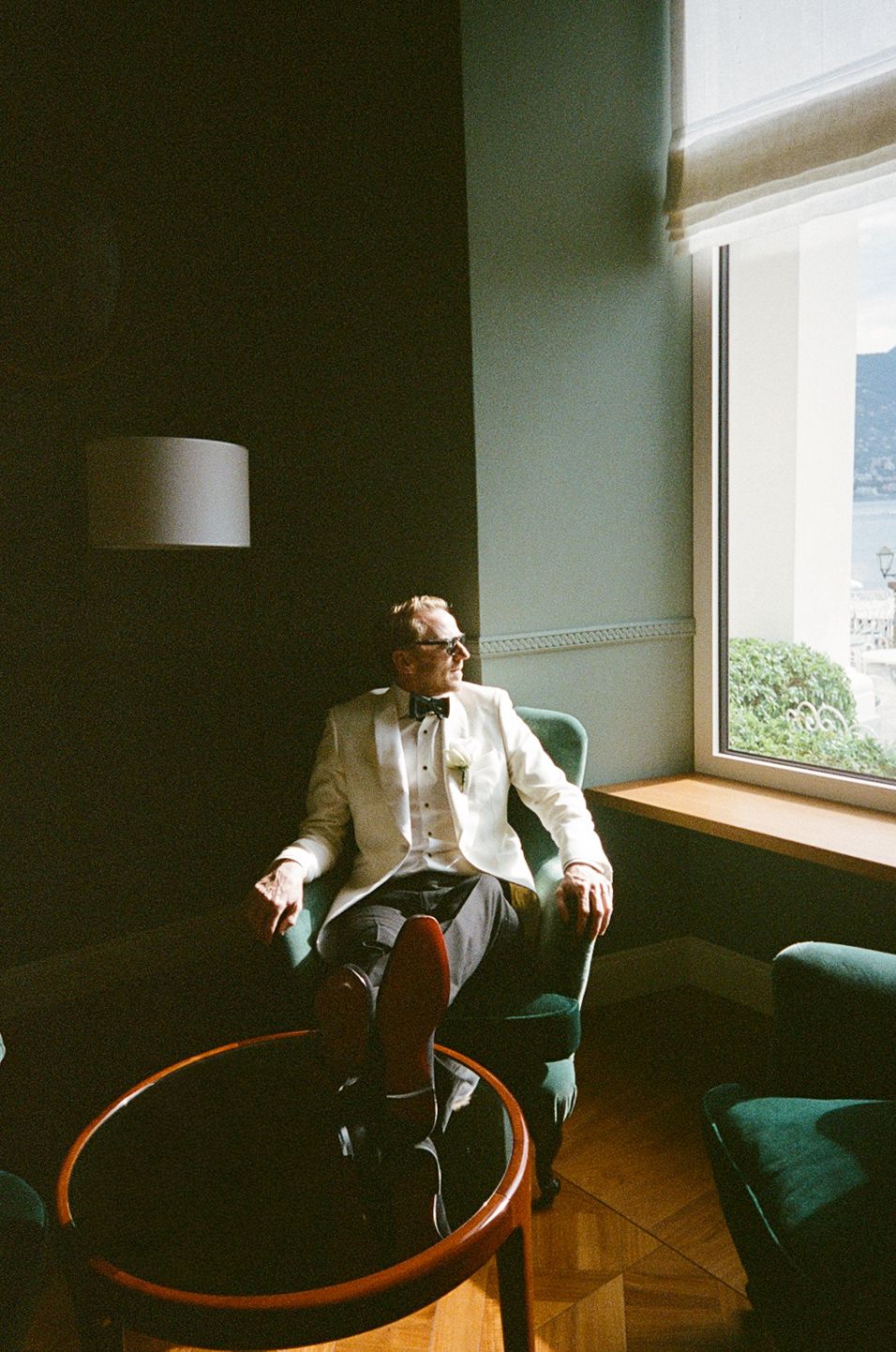 A dapper man in a white blazer and bow tie sits in a green velvet chair by a sunlit window.