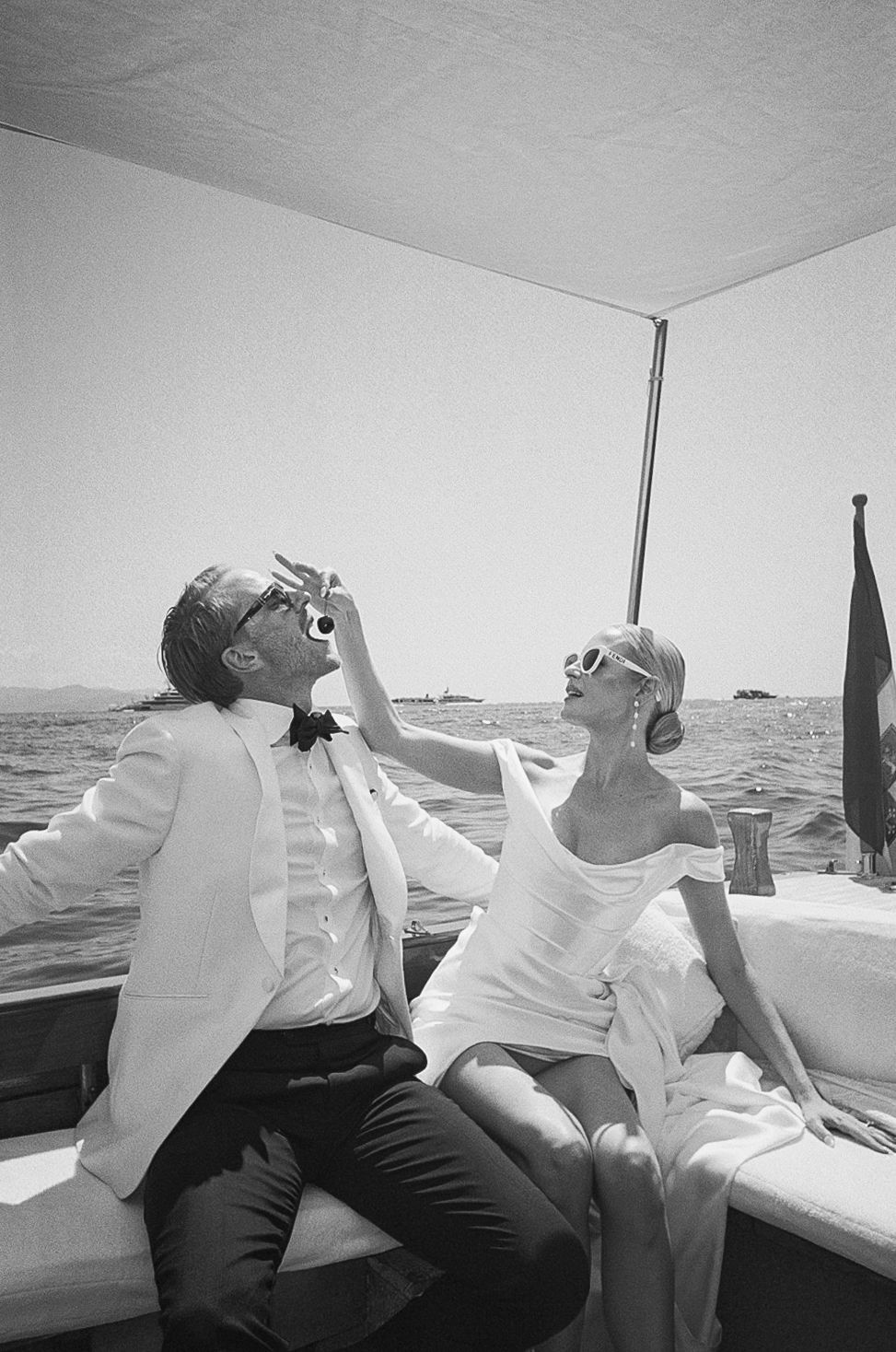 Newlyweds in white wedding attire laughing together on boat deck, black and white photo.