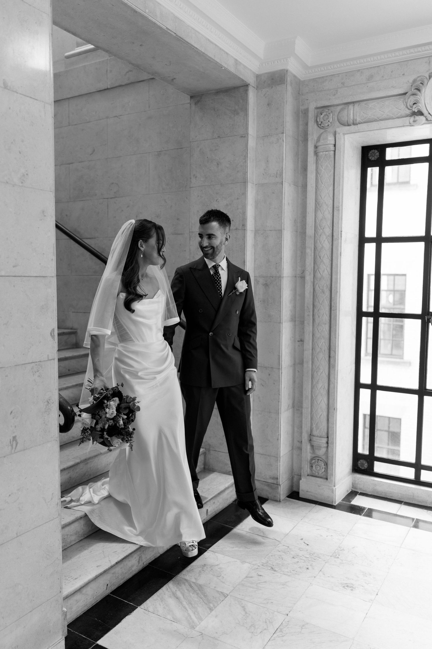 Black and white photo of smiling bride and groom on marble stairs at their wedding venue.