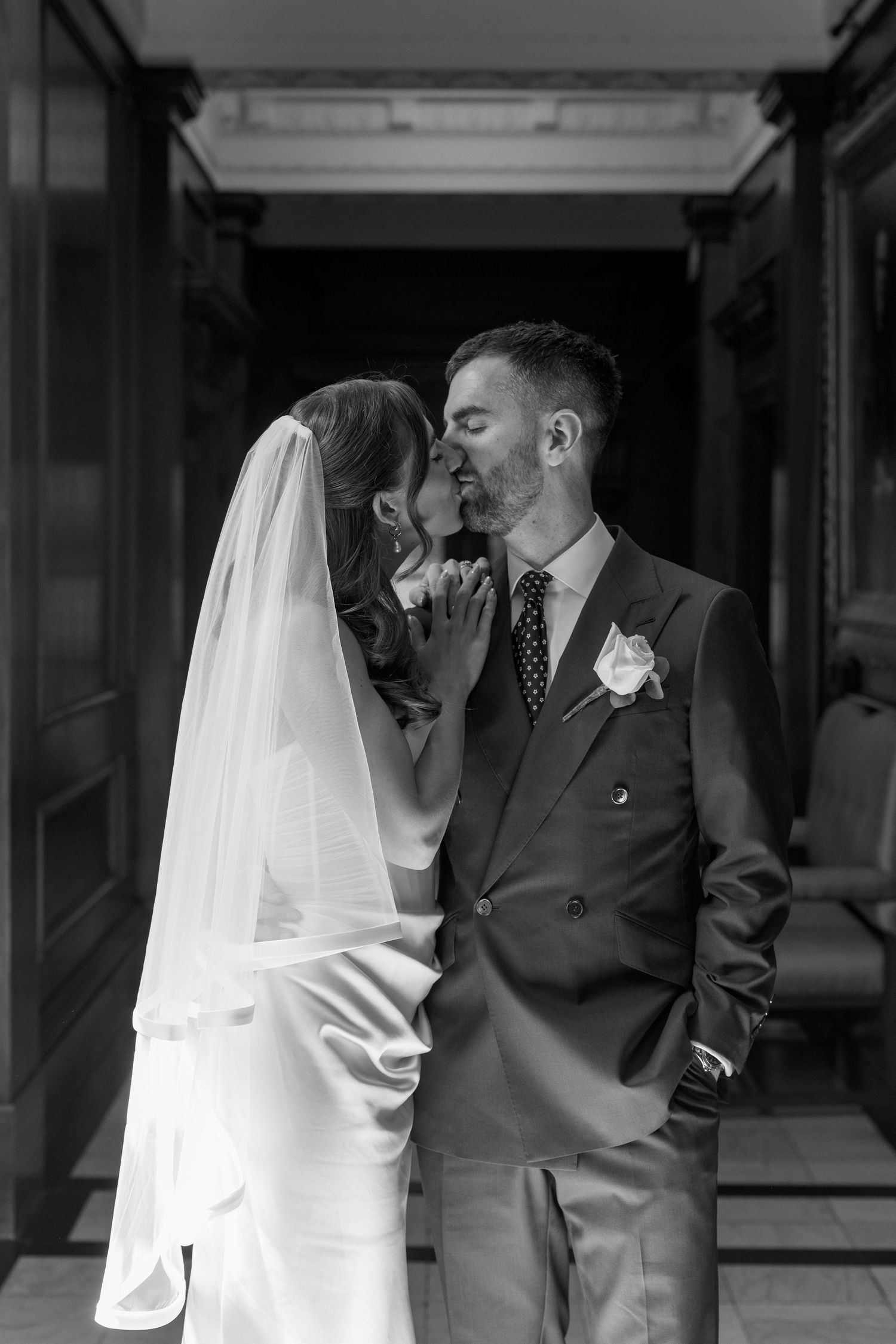 Black and white portrait of bride and groom sharing a kiss in elegant columned hallway.