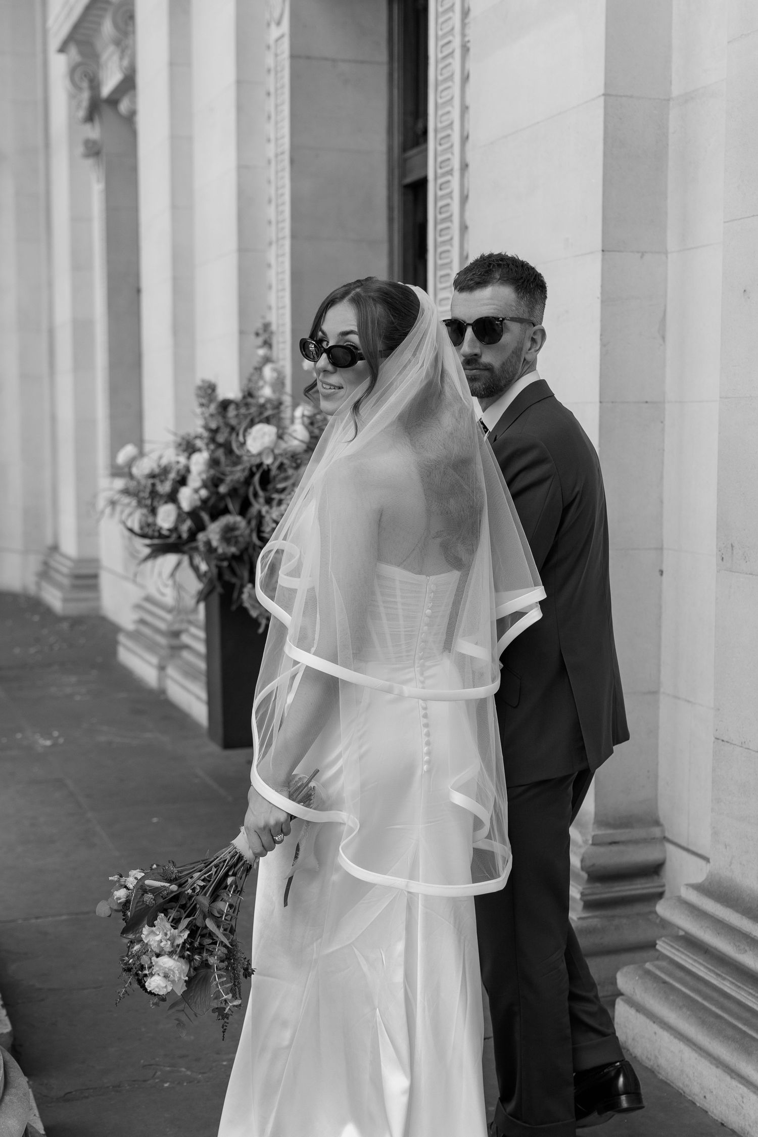 Black and white photo of bride in veil and sunglasses with groom beside classical stone columns of elegant building.