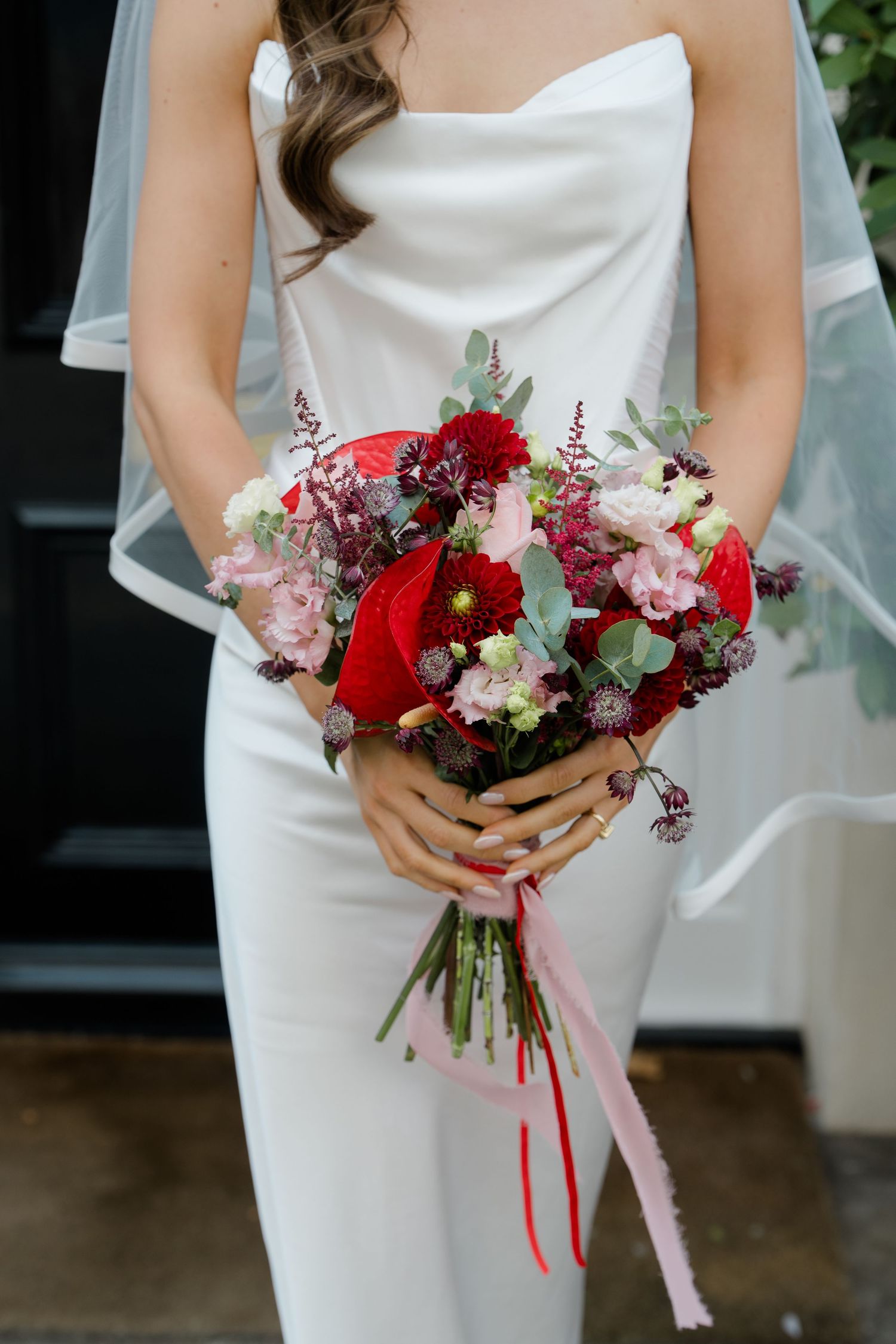 Bride in white strapless gown holds vibrant red and pink wedding bouquet with ribbon streamers.