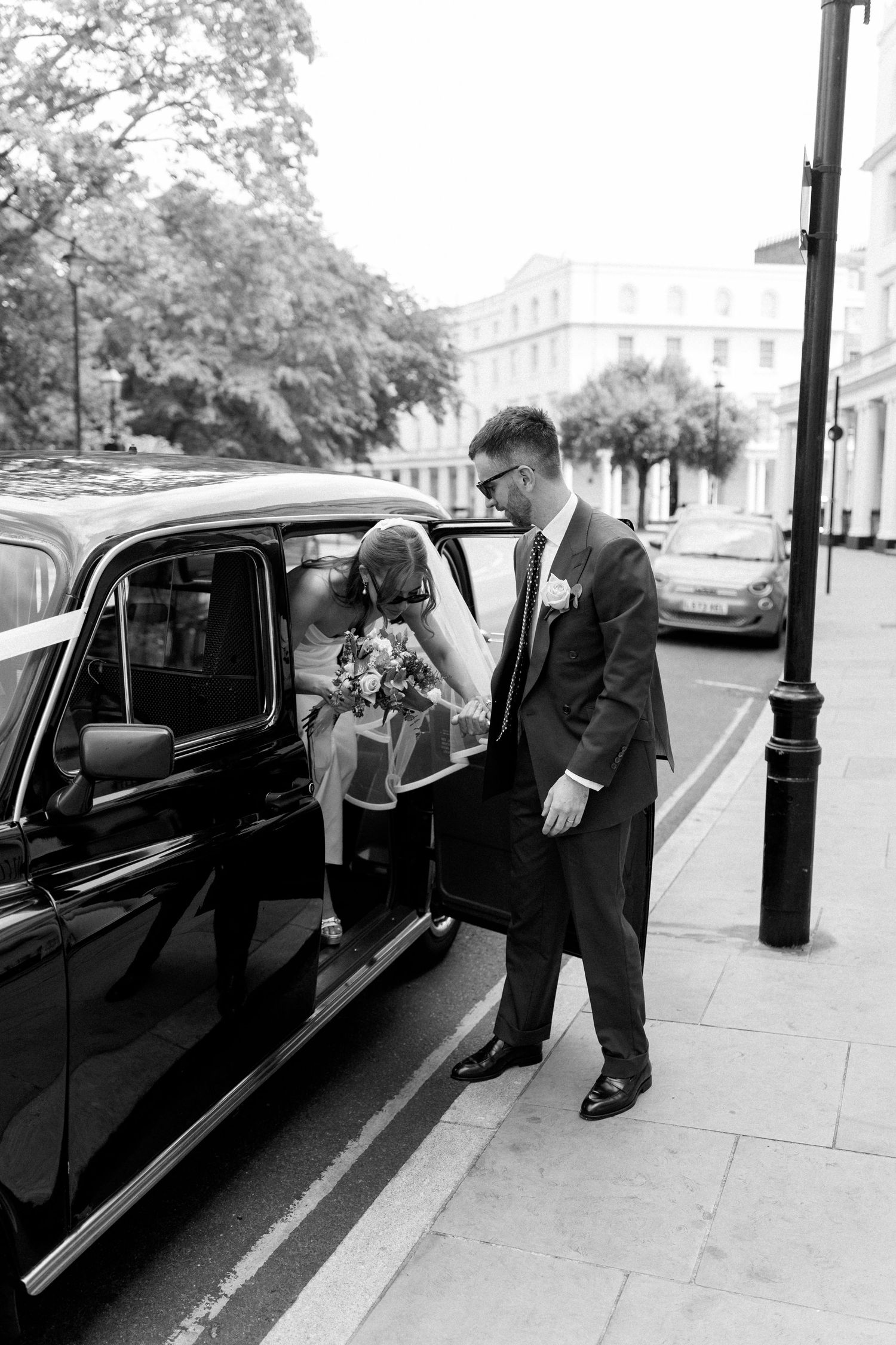 Black and white photo of groom in sunglasses helping bride exit a classic black London taxi.