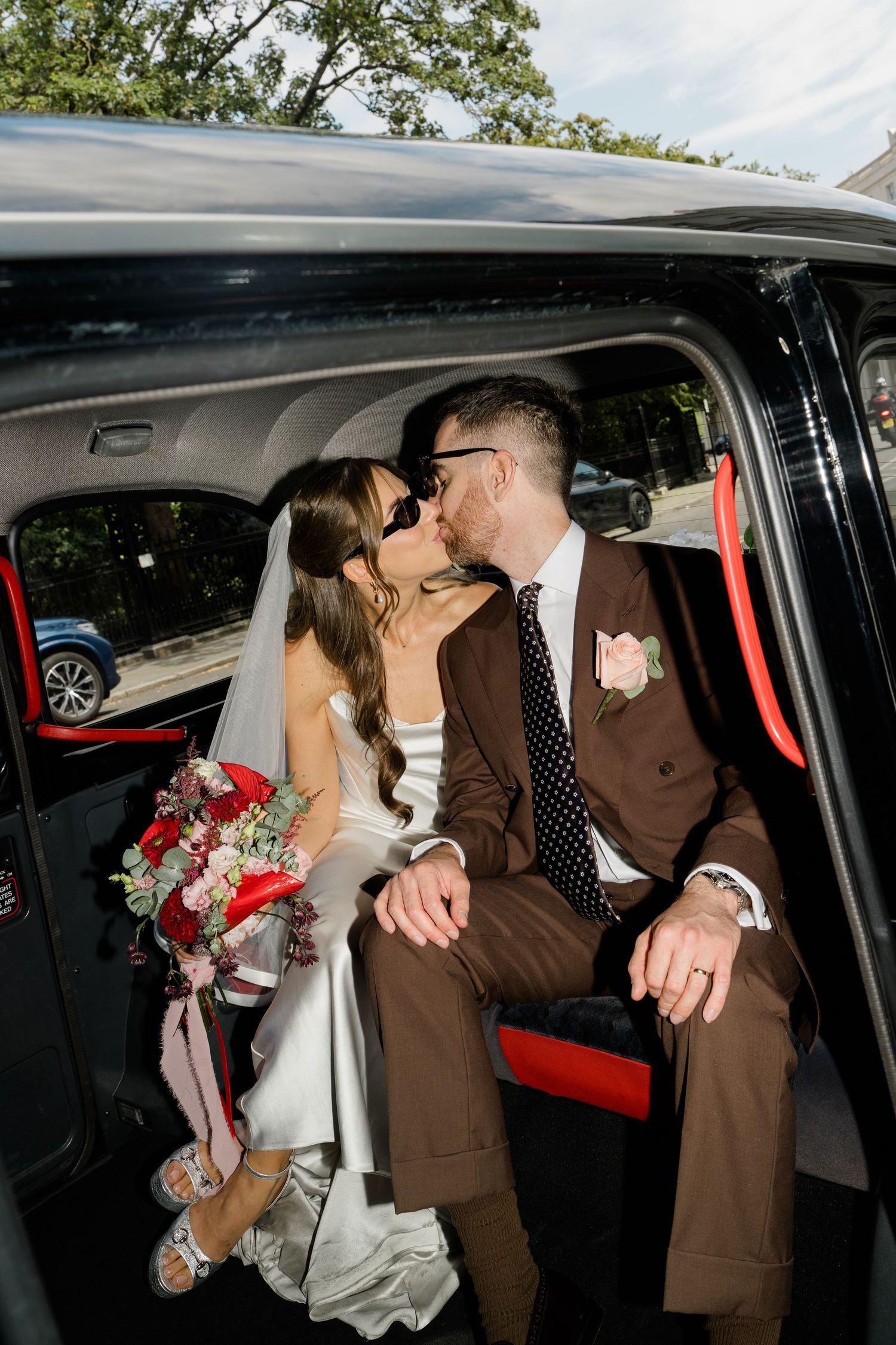 Bride and groom kiss inside black London taxi cab, bride holding red and white wedding bouquet.