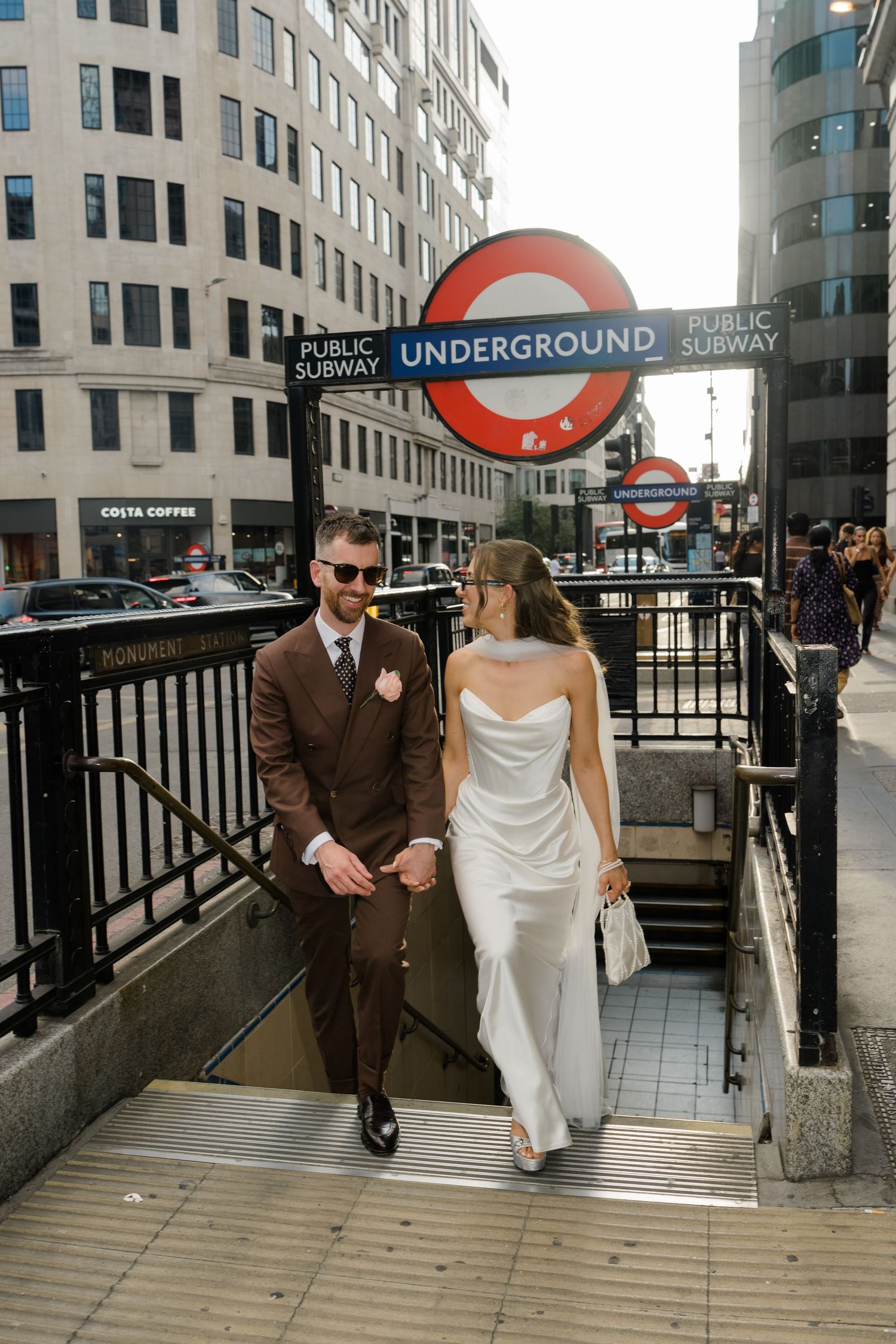 Bride and groom emerge from London Underground Monument station in wedding attire on sunny day.