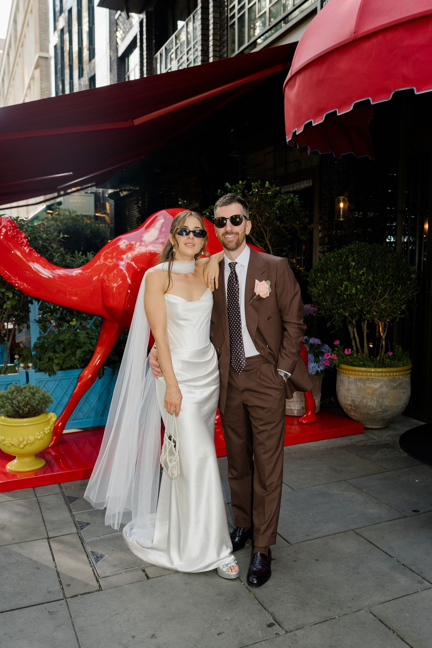 Bride in white satin gown and veil poses with groom in brown suit outside red awning restaurant.