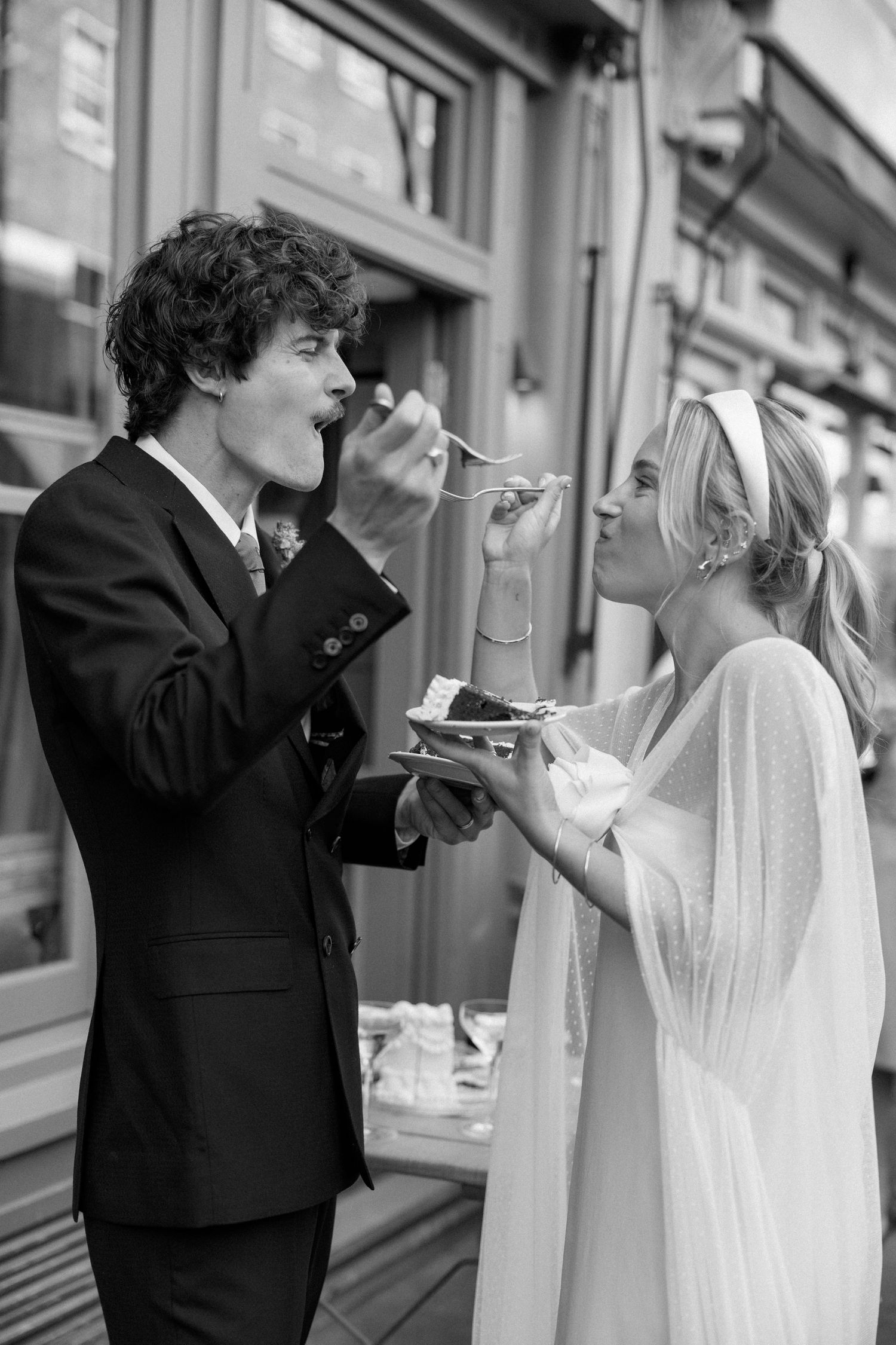 Wedding couple shares cake bites in a series of black and white photos outside a venue.