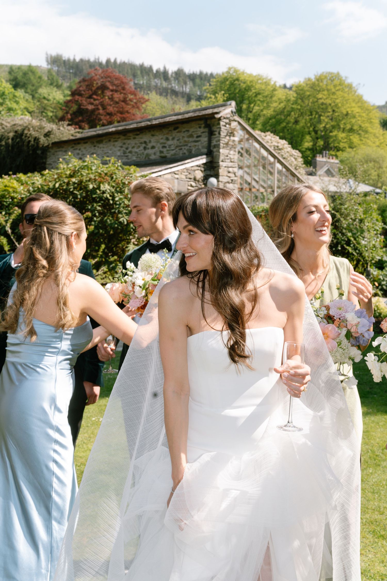 Wedding party in pastel dresses celebrates on lawn with stone building and mountain views.