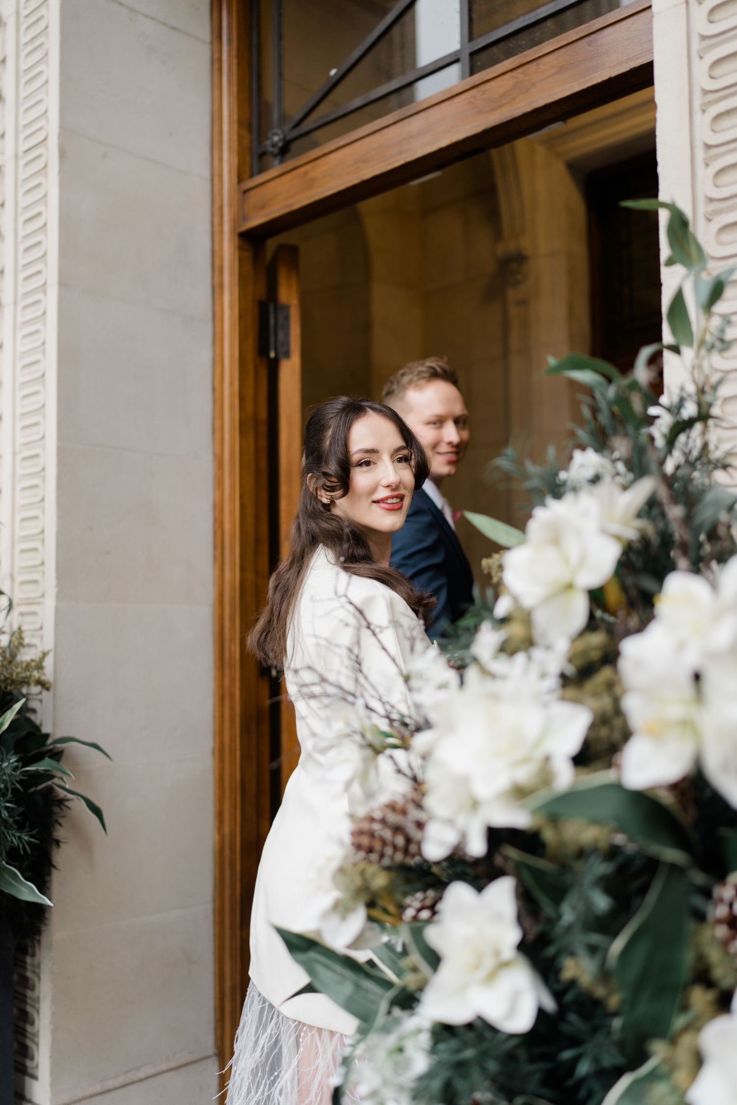 Bride in white blazer and feather skirt smiles at camera beside groom at elegant wooden doorway with floral arrangement.