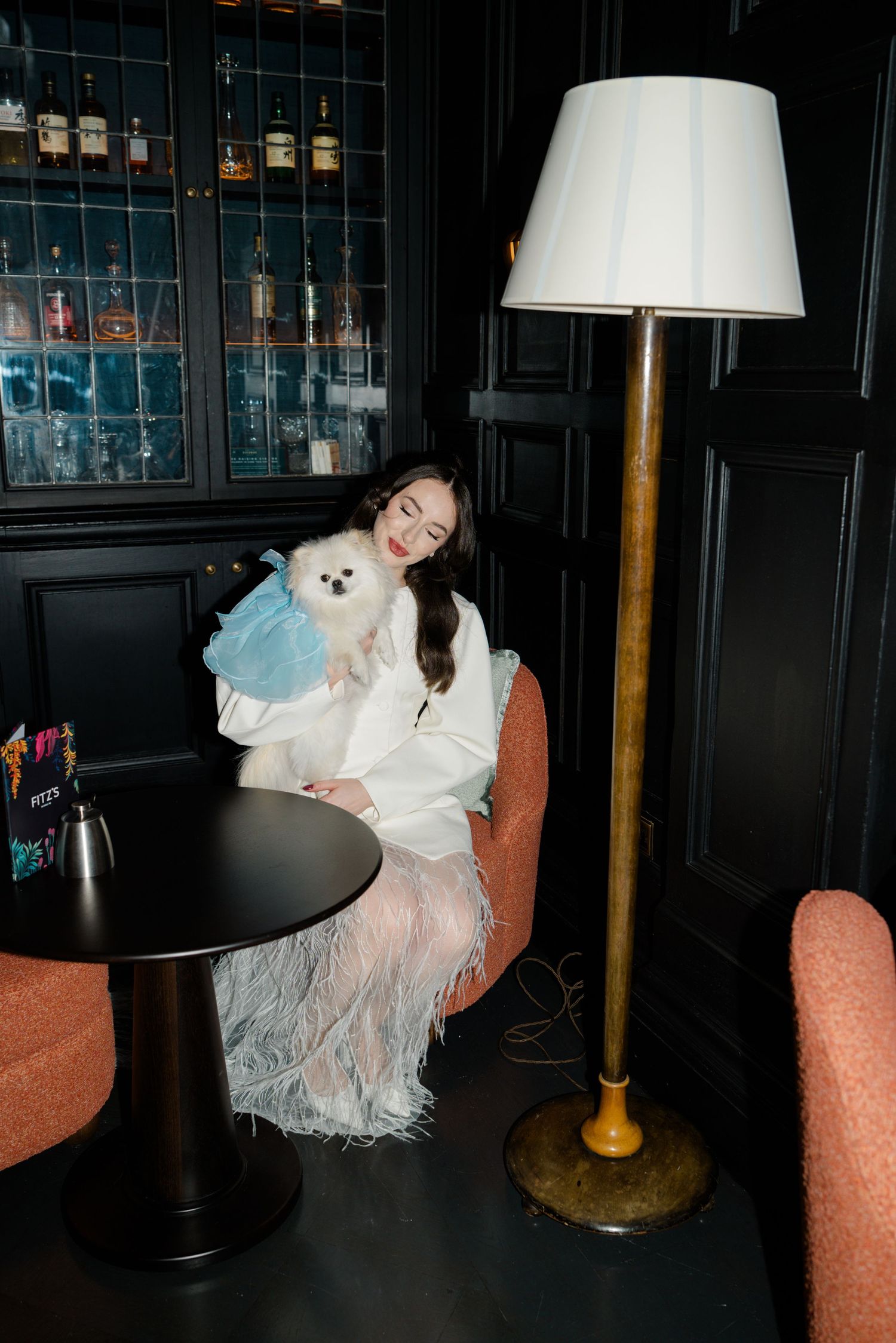 Elegant bride in white feather skirt holding small white Pomeranian dog in blue dress at dark bar.