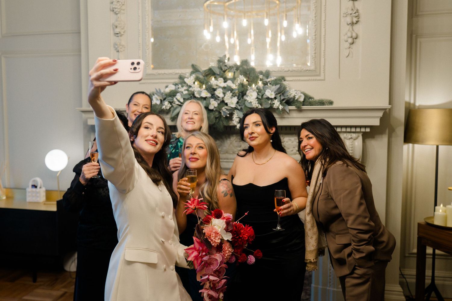 Group of five women taking a selfie at a wedding reception near floral fireplace.