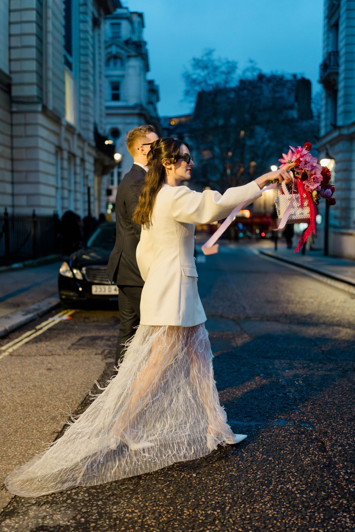 Bride in white feathered dress holding bouquet on London street at dusk with groom behind her.