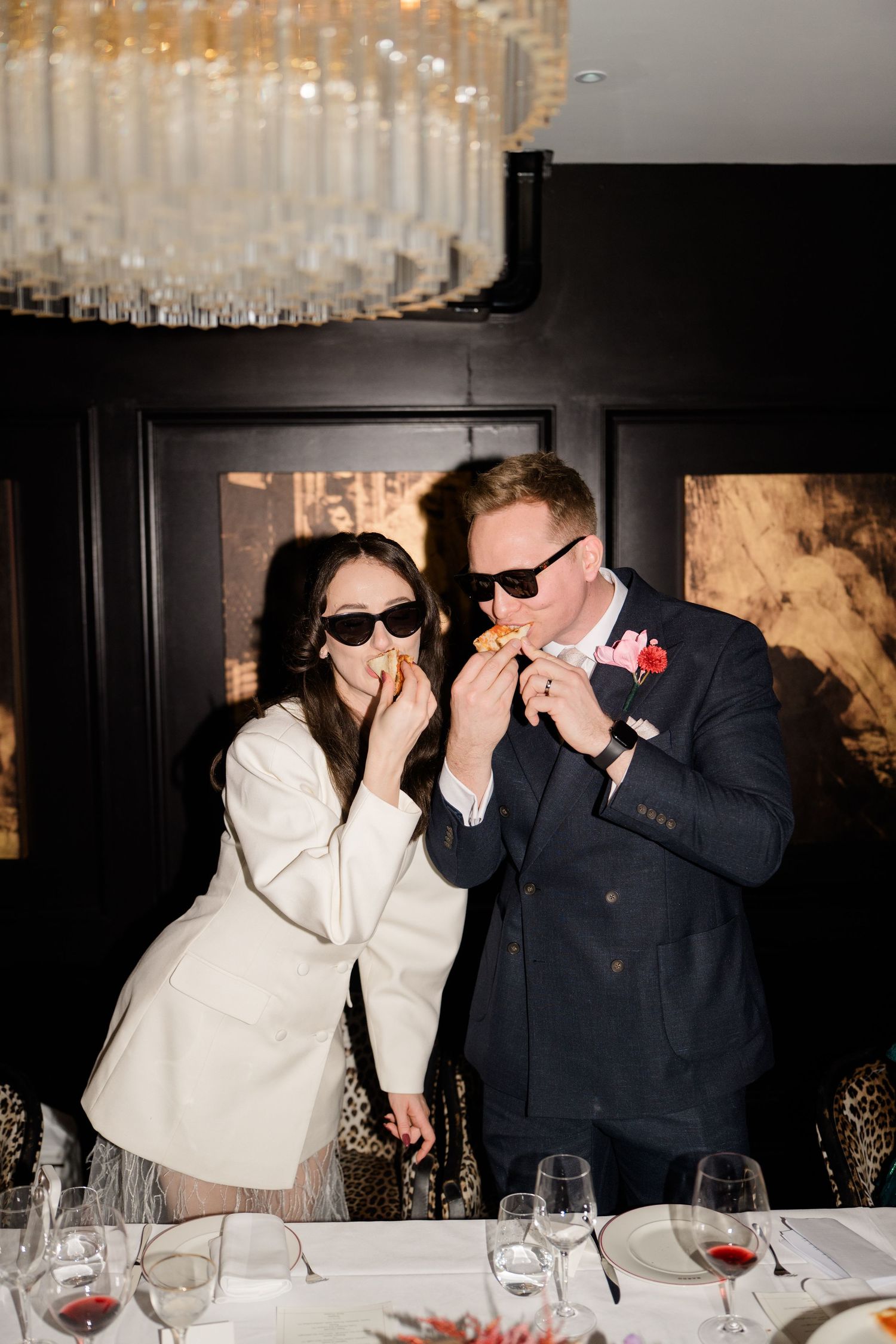 Bride and groom wearing sunglasses playfully eat appetizers together at elegant wedding reception dinner table.
