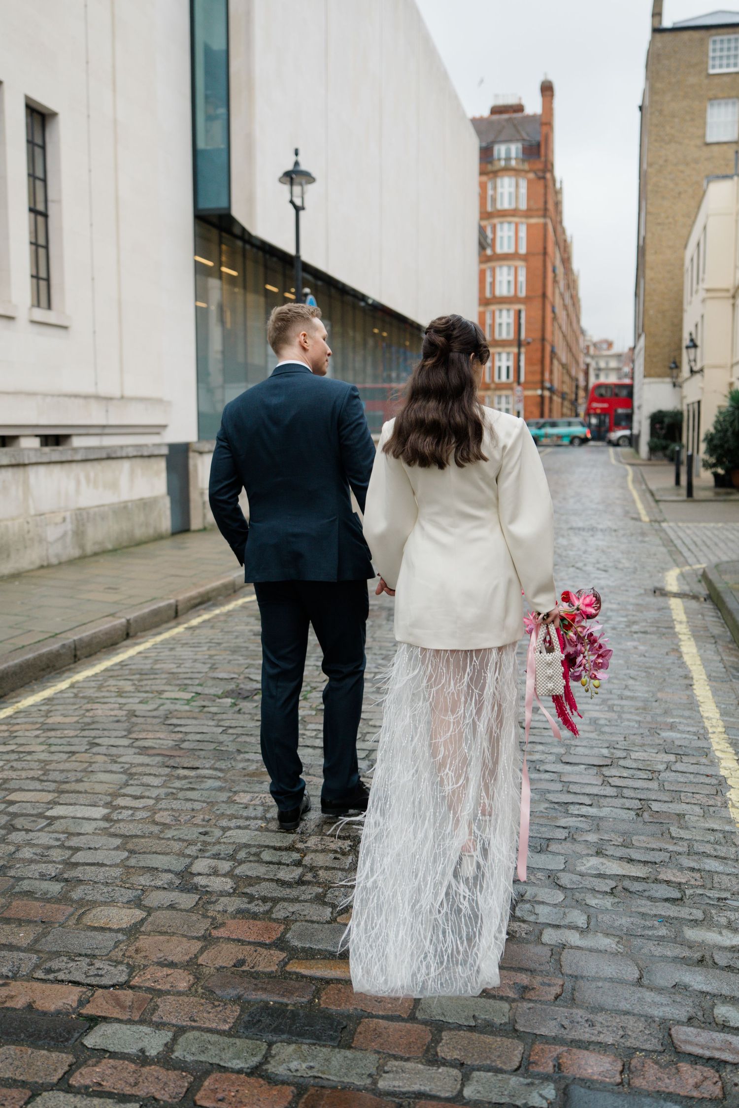 Bride in white feather skirt and groom in navy suit walk cobblestone London street holding hands.