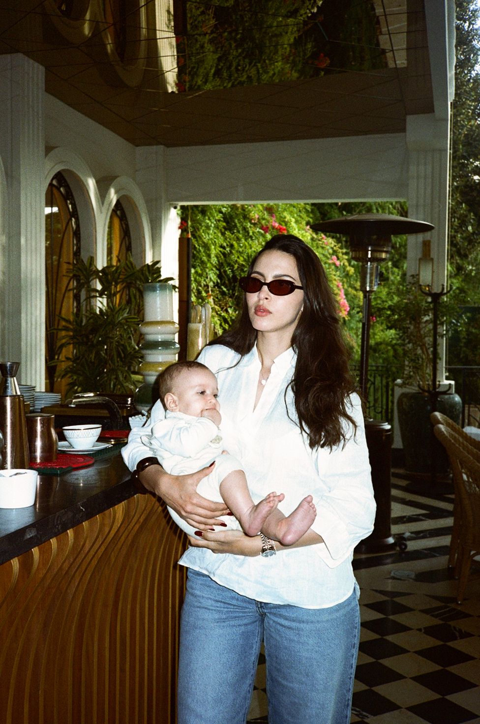 Young woman in white shirt and jeans holding a baby, wearing red sunglasses in an elegant restaurant.
